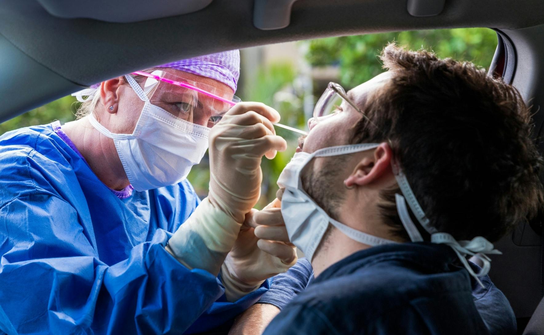 A female health worker wearing blue scrubs, a white mask and eye protection, leans through a car window to swab a man sitting in a front seat.