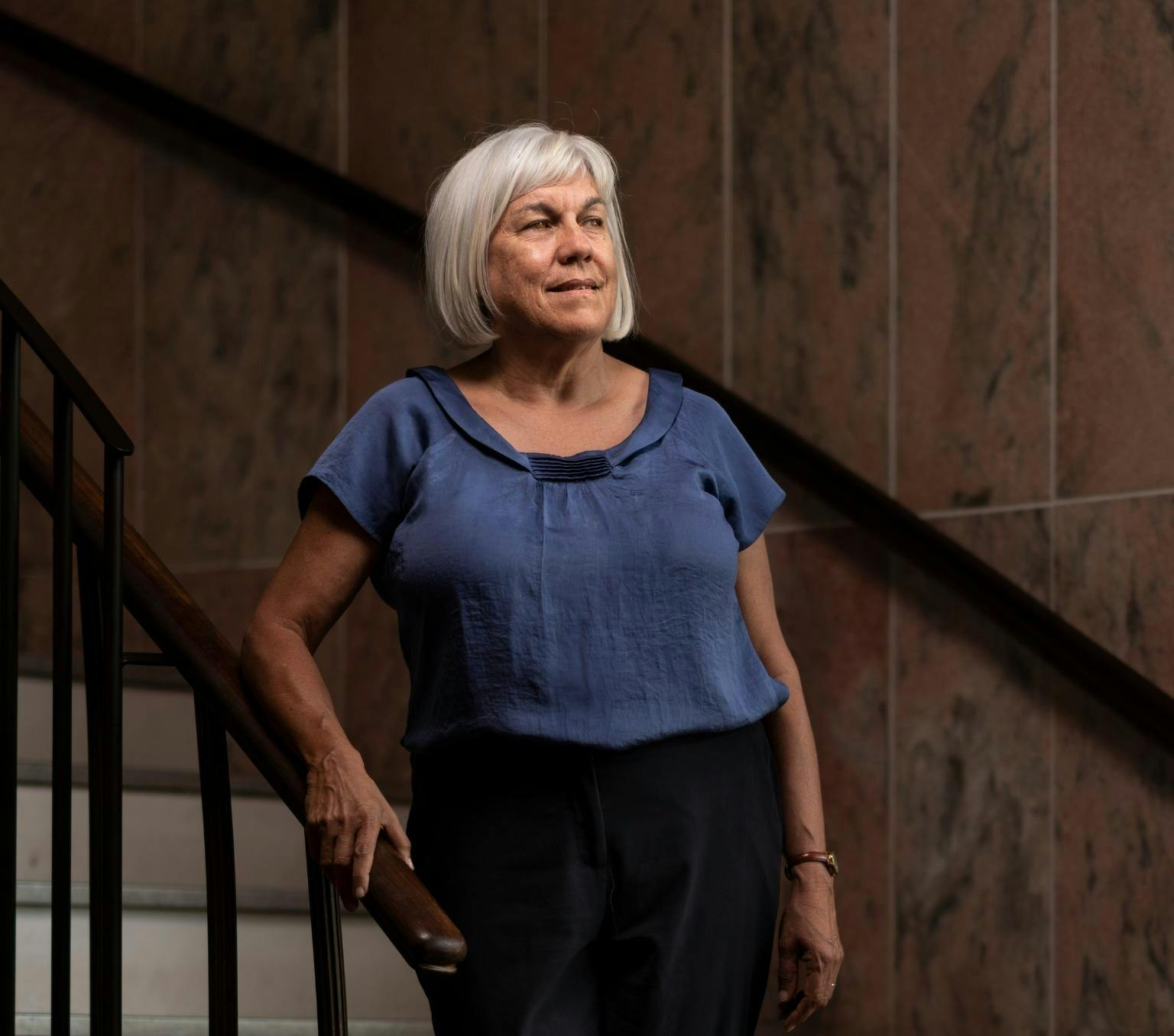 Jill is wearing a blue blouse and black pants. She has a grey bob. She is standing on a staircase in front of a brown stone wall and looking past the camera