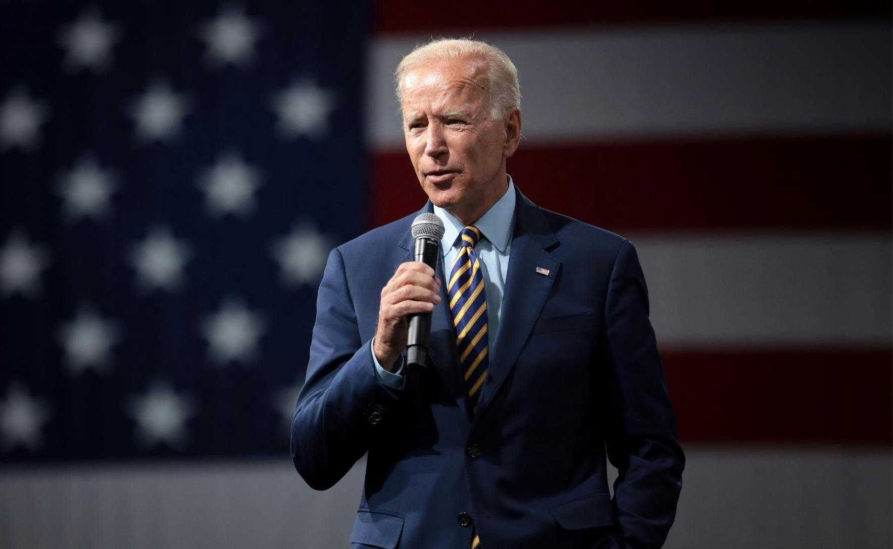 Jo Biden is wearing a dark blue suit and yellow and blue striped tie. He is holding a microphone and mid speech. Behind him is the US flag