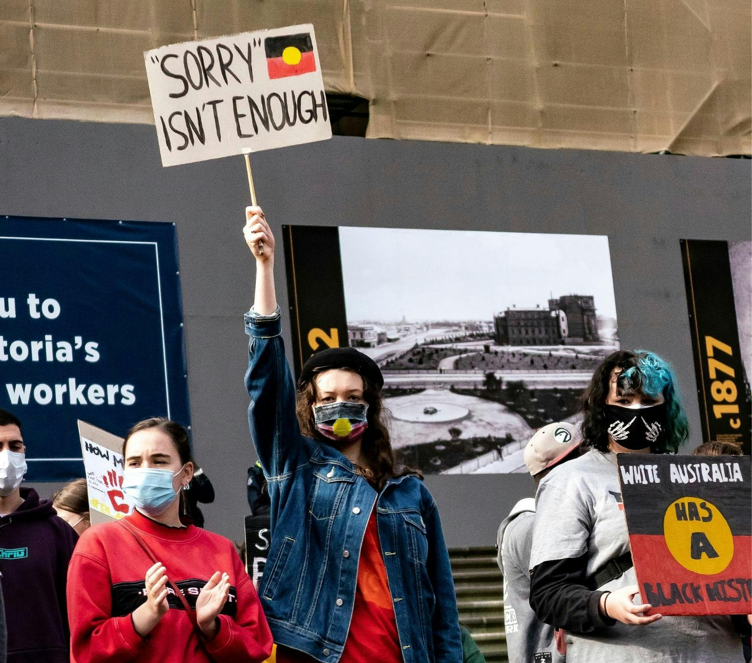 People at a Black Lives Matter protest in Australia. People are wearing red yellow and black and masks. One person is holding a sign that reads "sorry isn't enough".