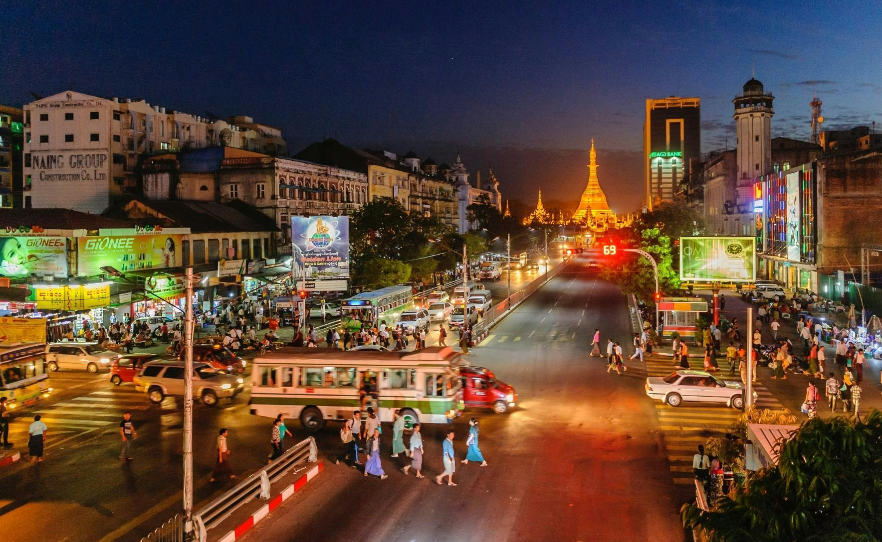 Myanmar street at night. A gold temple is in the background. there are buildings with bright lights and signs lining the streets and cars on the road