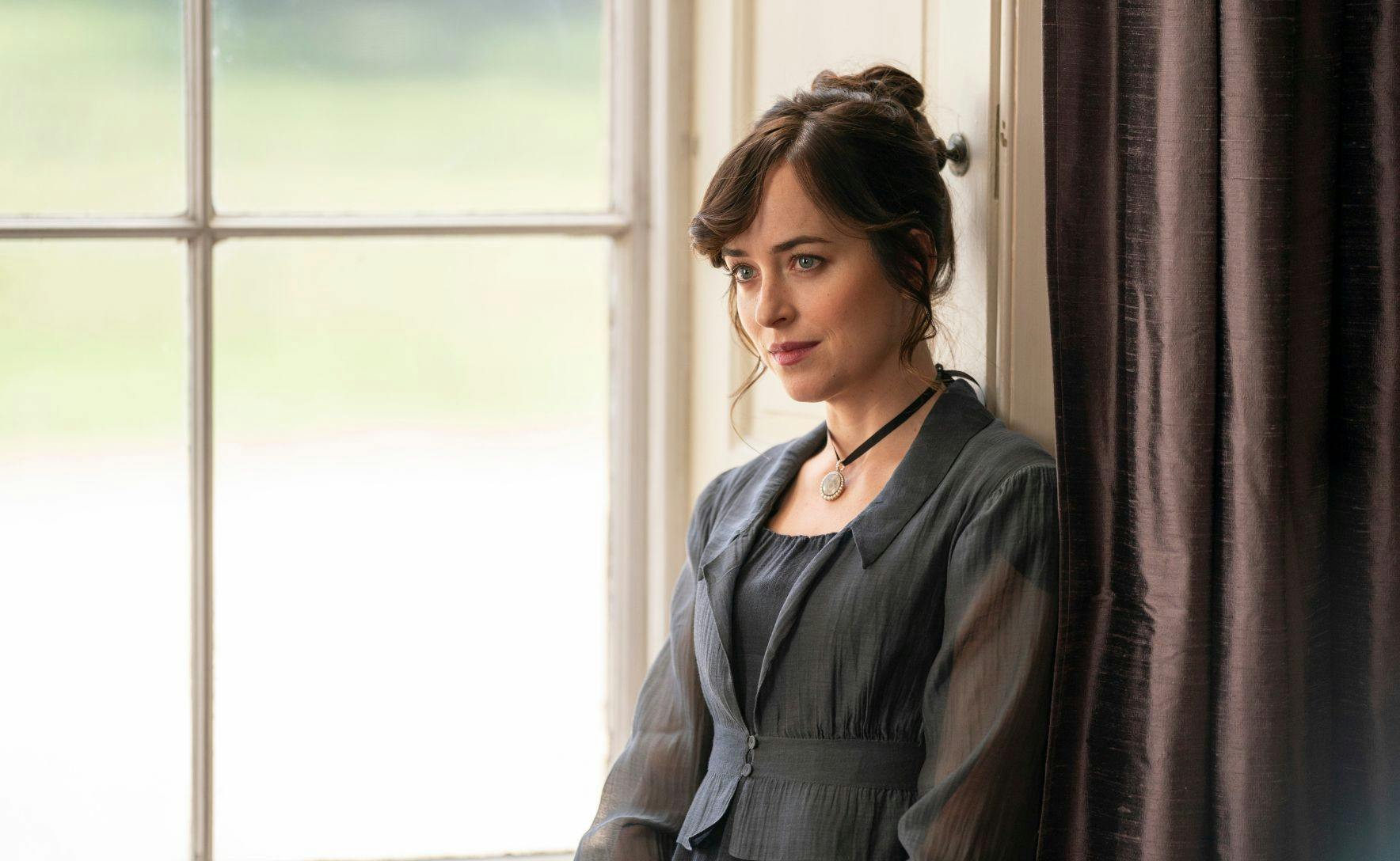 Dakota Johnson as Anne Elliot in Persuasion. She is leaning against a window, with brown curtains behind her. She wears a grey Regency style dress and her reddish hair is in a messy bun.