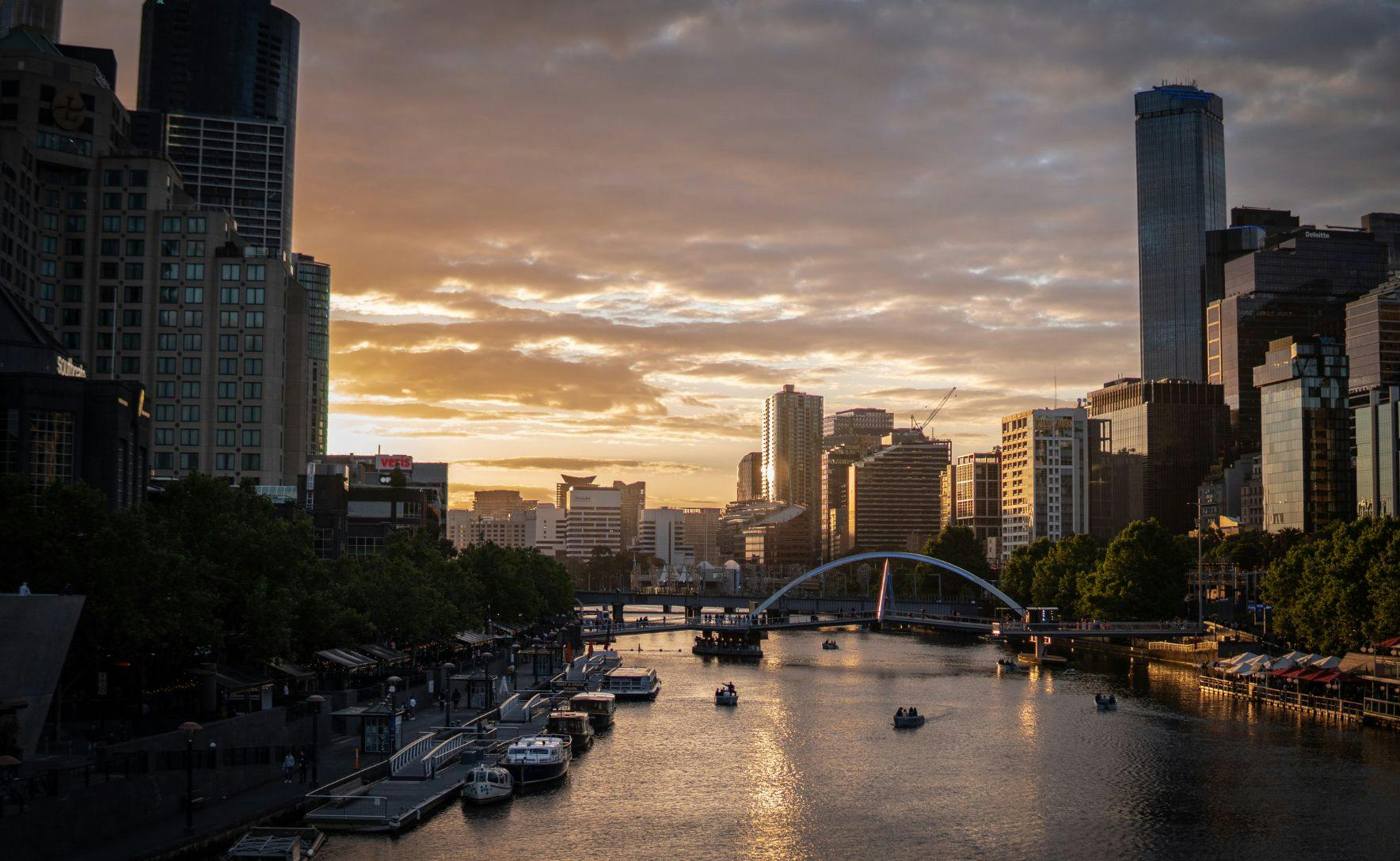 A shot of the Yarra river in Melbourne, with city buildings along both sides. There are boats in the river, and the sky is the orange of a sunset.