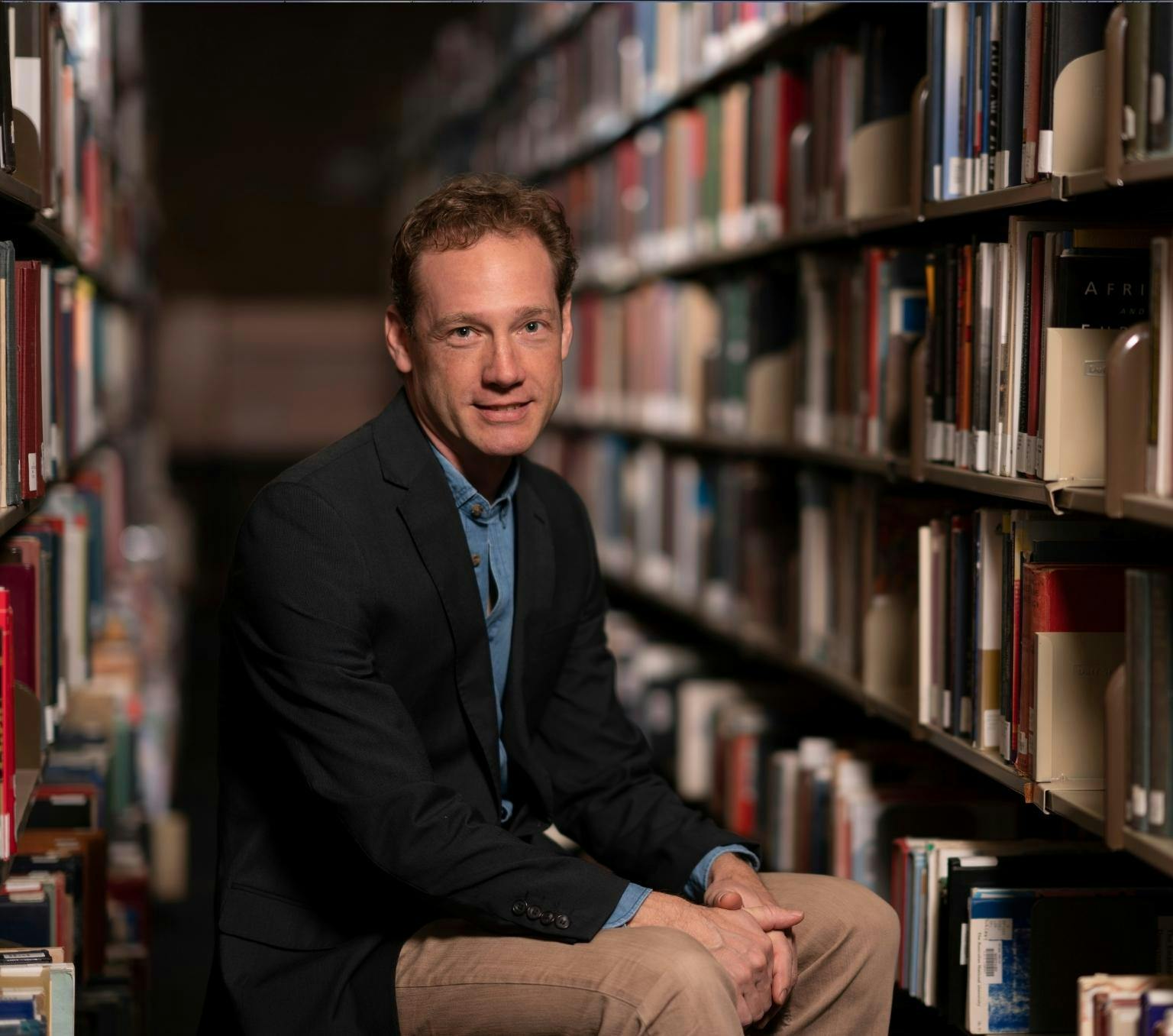 Timo is sitting in an aisle of a libary surrounded by books. He is looking at the camera with a netral expression and wearing a black suit jacket, beige pants and blue collared shirt.