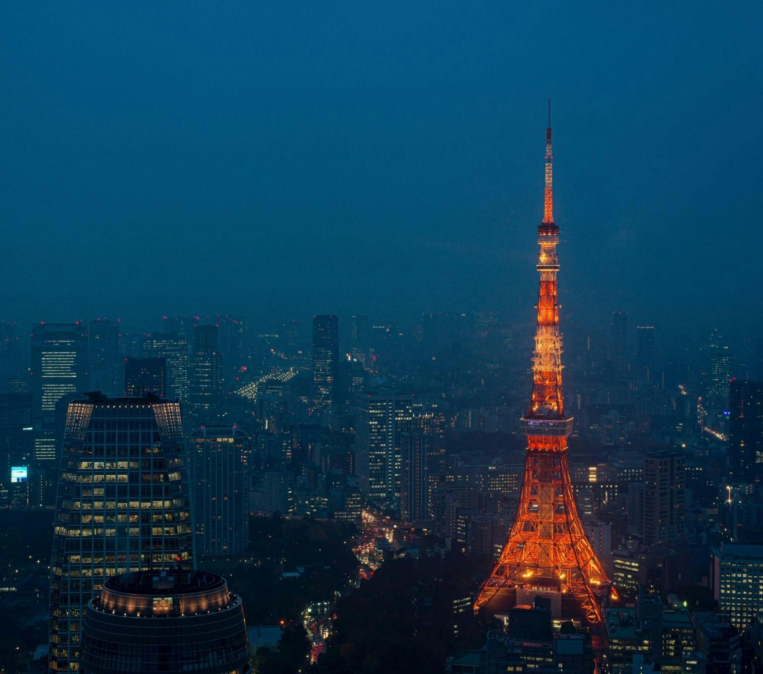 Tokyo tower which is red and lit with orange lights is pictured above the tokyo skyline at dusk