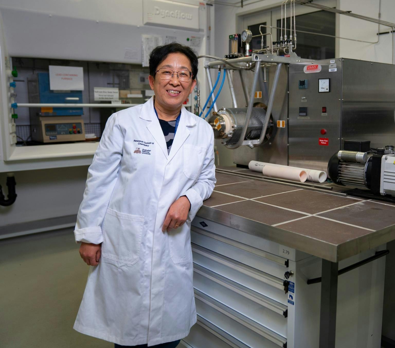 Yun Liu is wearing a white lab coat and smiling at the camera. She is standing in a laboratory