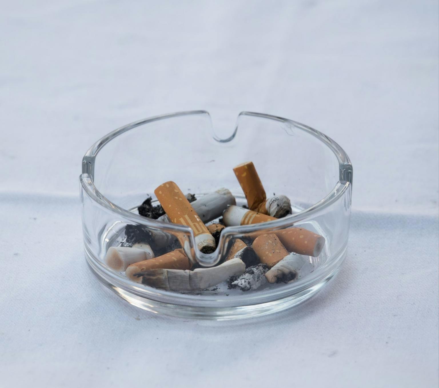 glass ashtray on a white table cloth. the ashtray is filled with burnt cigarettes and ash