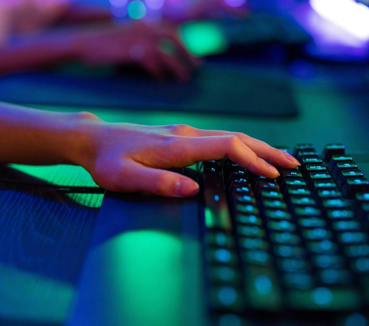 close-up of a persons hand resting on a keyboard. Green and blue light illuminates a blurred background and the keyboard