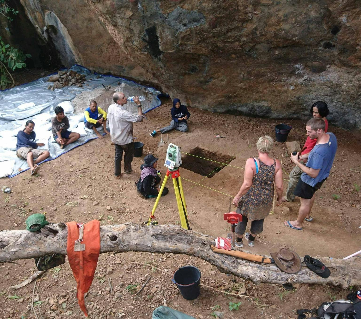 excavation site. four people are standing around a square hole cut into the brown earth. Three people are sitting on a large silver sheet of plastic. There is a large brown rock face in the background and leaves. Various excavation equipment is laying around the site