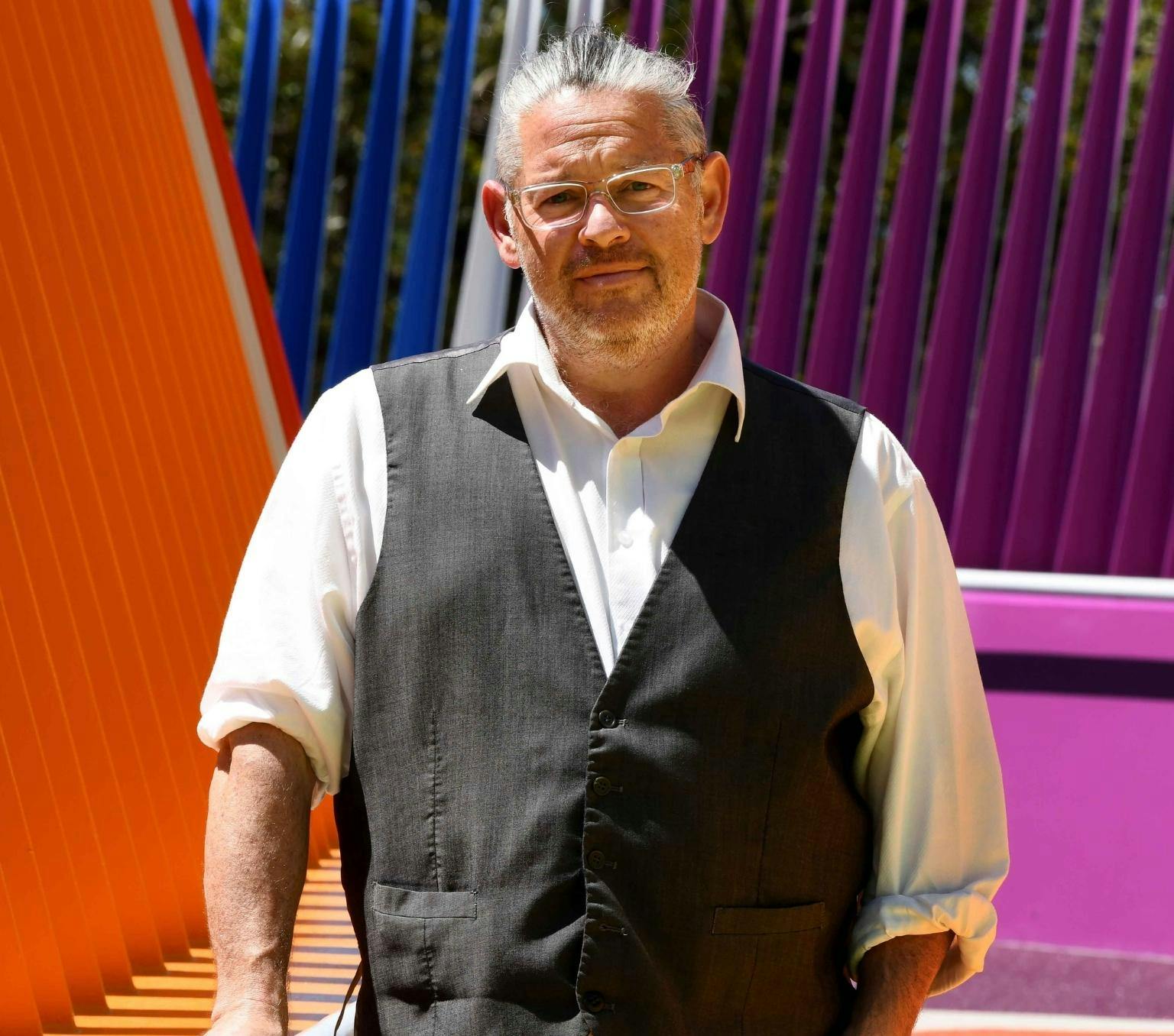 Alex Brown is standing in front of an orange, purple and blue background. He is wearing a white collared shirt and a grey vest.