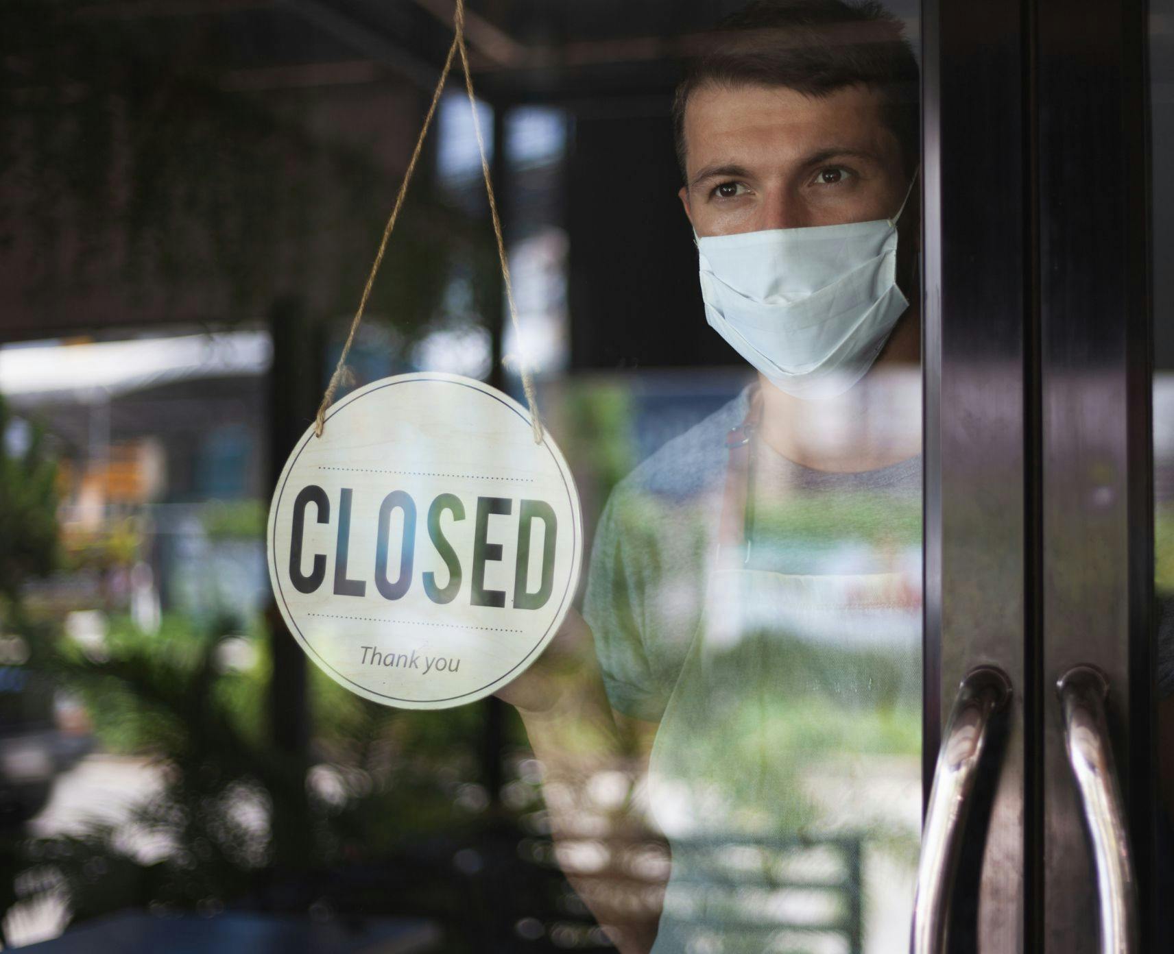Man at door of cafe wearing mask behind closed sign.