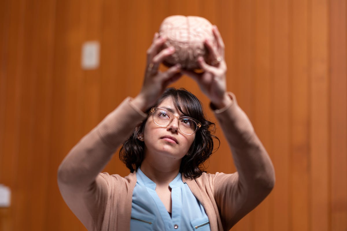 Daniela is holding a model of a brain above her head and looking at it. She is in a room with wooden panel walls.