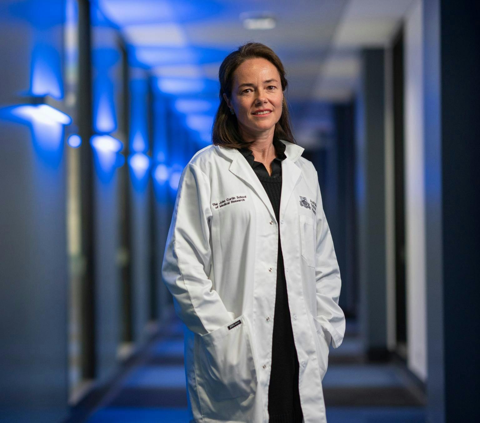 Carola is pictured in a corridor illuminated by blue lights. She is looking at the camera and wearing a white lab coat