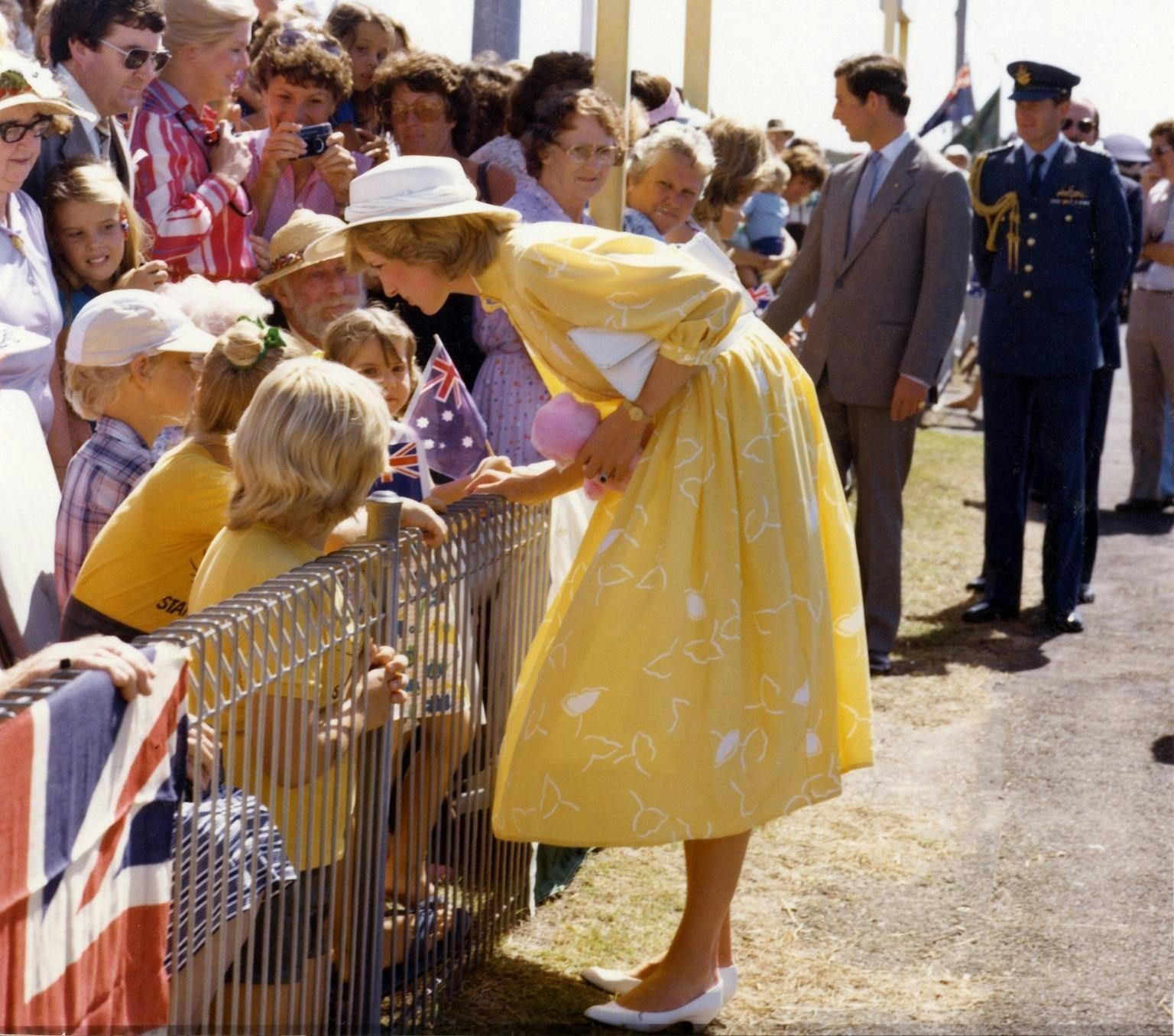 Diana is wearing a yellow dress and white hat. She is outside and leaning over a fence talking to children while crowds of adults watch on. Prince Charles is in the background also taking to crowds behind a fence