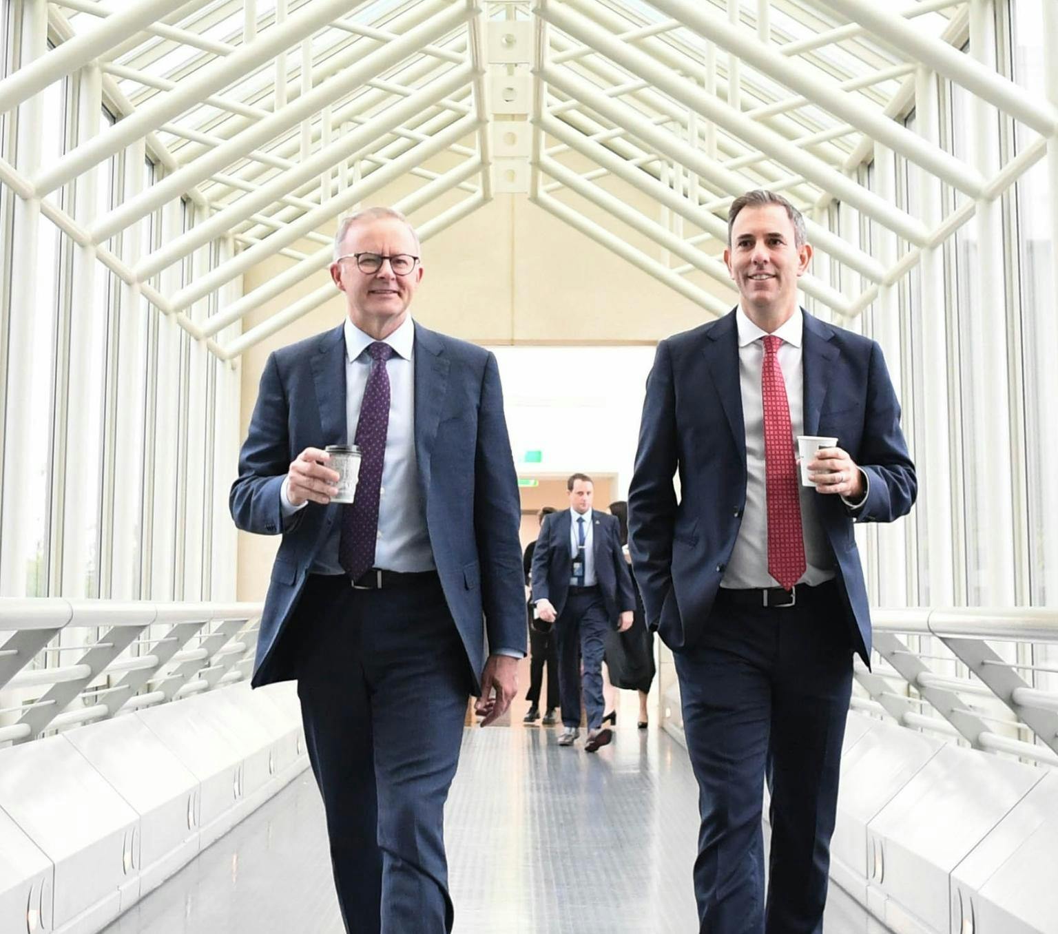 Two men in suits walk down a hallway that has big glass windows and a glass roof.