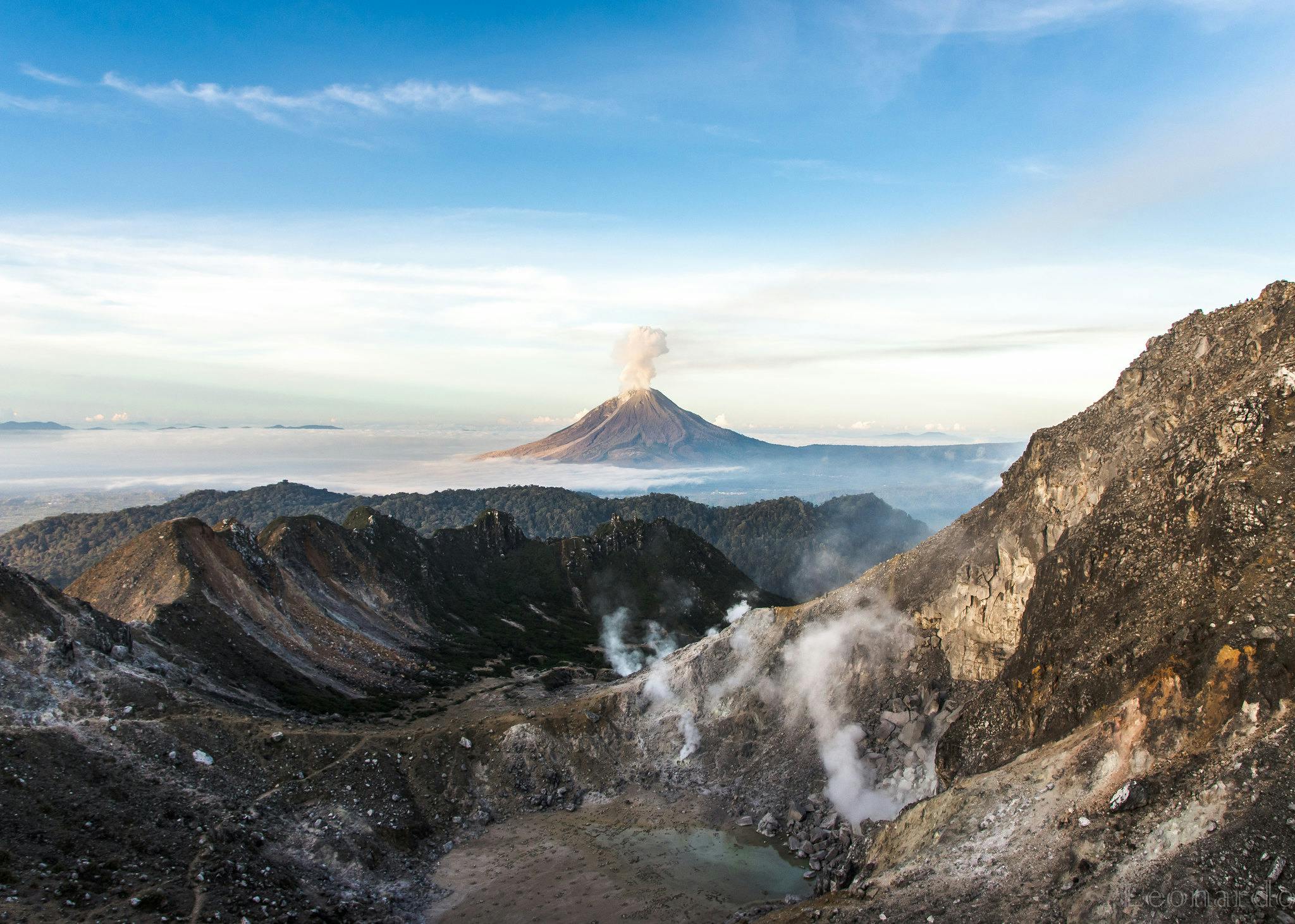 a volcano is visible sitting above clouds in the background. In the foreground are rocky hills and craters