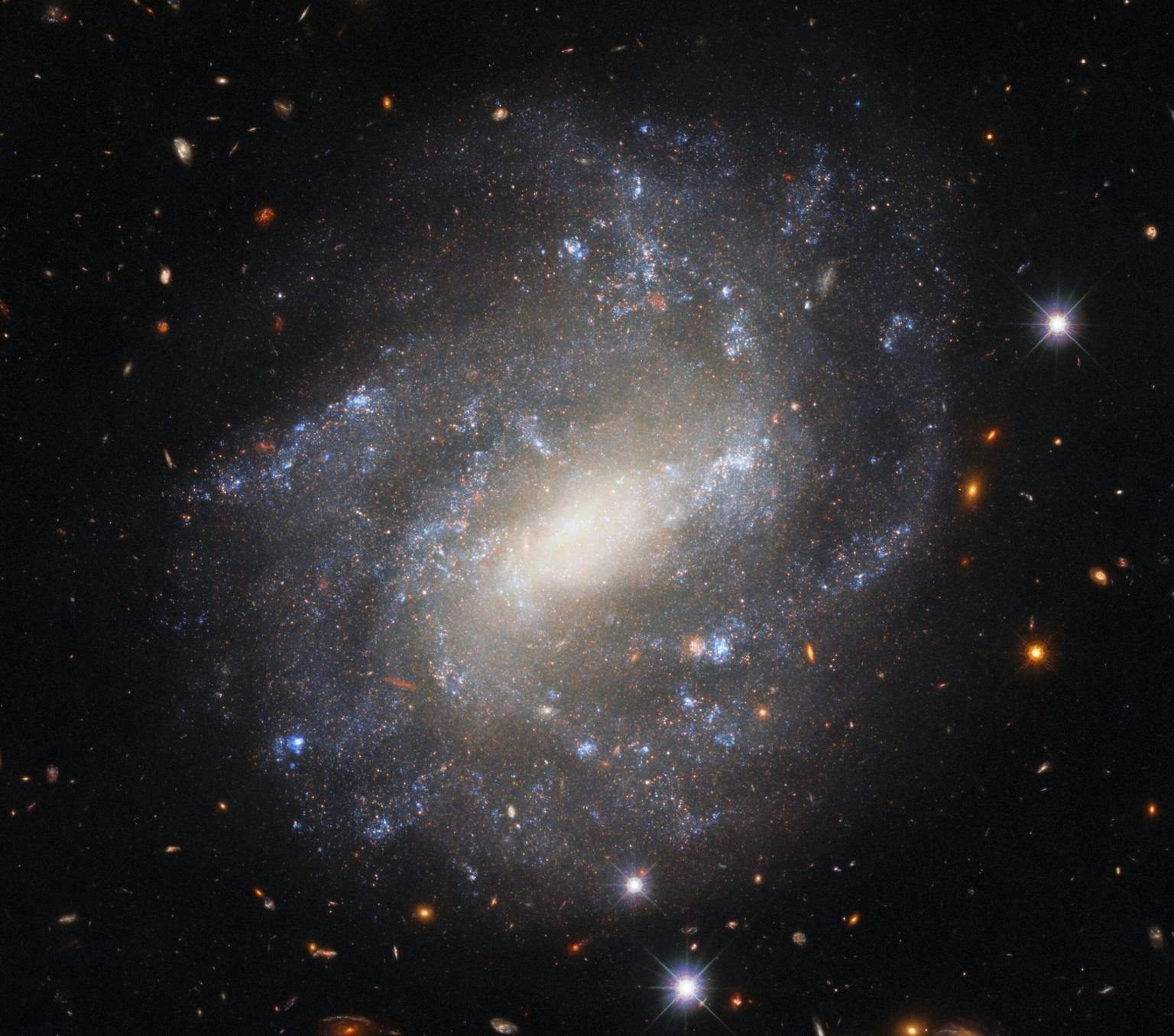 Image of a spiral galaxy. Its star-studded spiral arms stand in splendid isolation against a backdrop of distant galaxies, which are only visible as indistinct swirls or smudges thanks to their vast distances from Earth. The image also features some much brighter foreground stars closer to home. These bright nearby stars are ringed with diffraction spikes – prominent spikes caused by light interacting with the inner workings of Hubble’s secondary mirror supports.