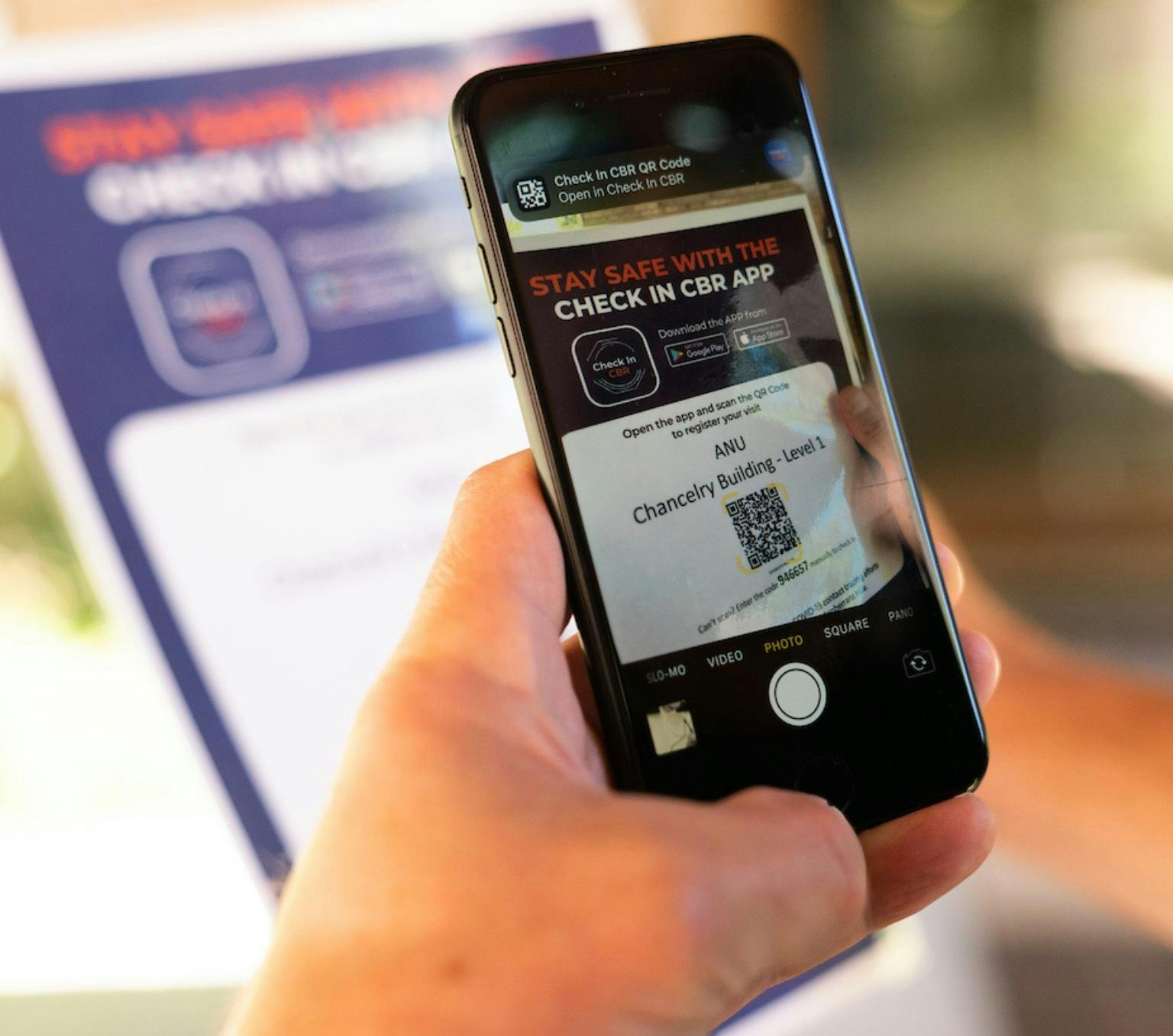 close up photo of a person's hand holding a phone infront of Canberra COVID-19 checkin posters. The phone displays the Canberra check-in app.