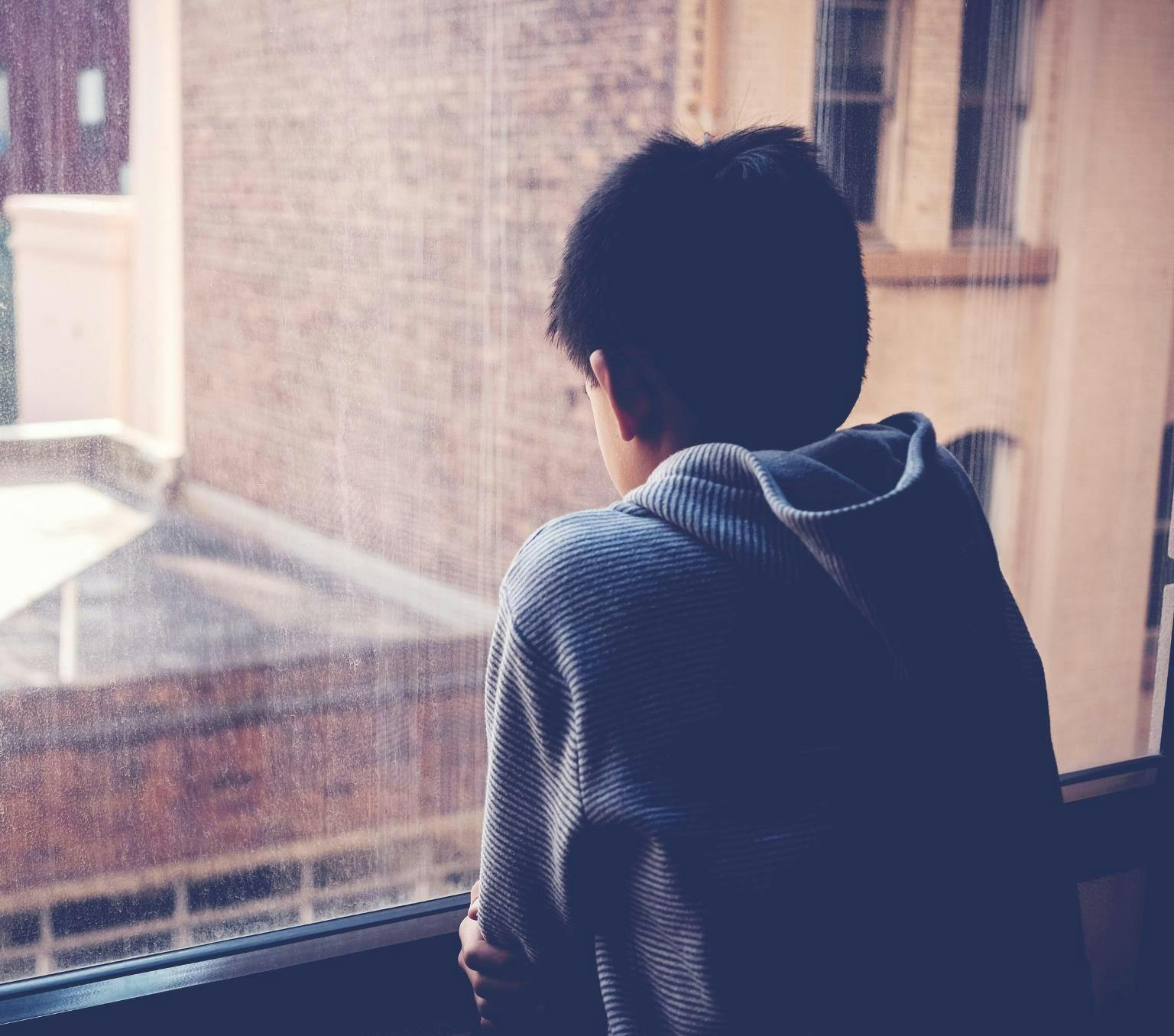 back of a boy with black hair looking out a windo onto houses with his shoulders hunched.