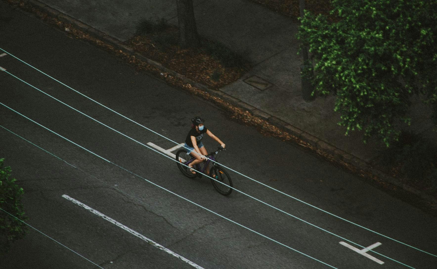 aerial shot of a woman riding a bike on a road. She is wearing a mask and a helmet. There is no one else in the image