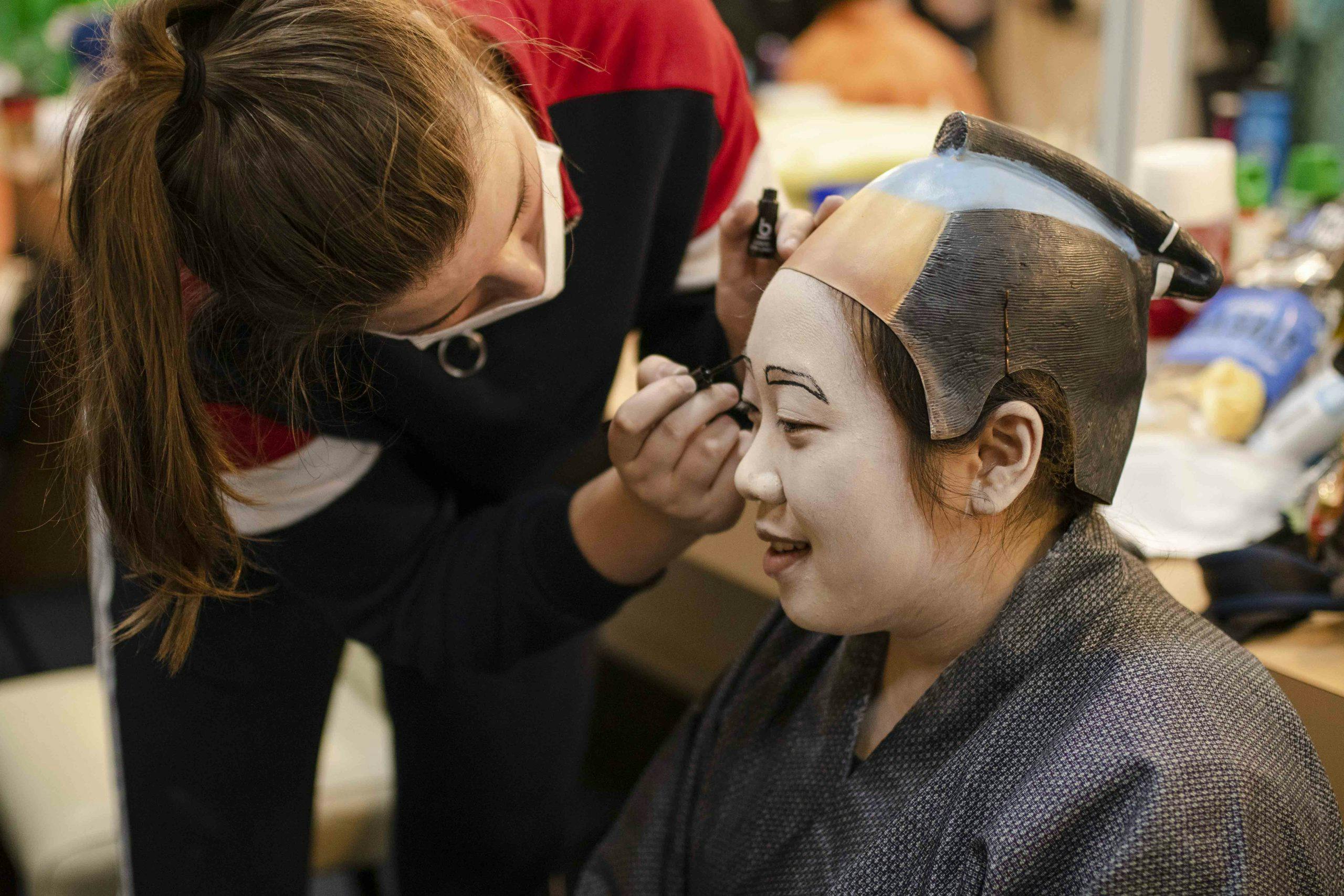 a woman in a mask is bending down and painting black eyebrows on a woman's face. The woman having her makeup done is wearing a kabuki costume and has her face painted white