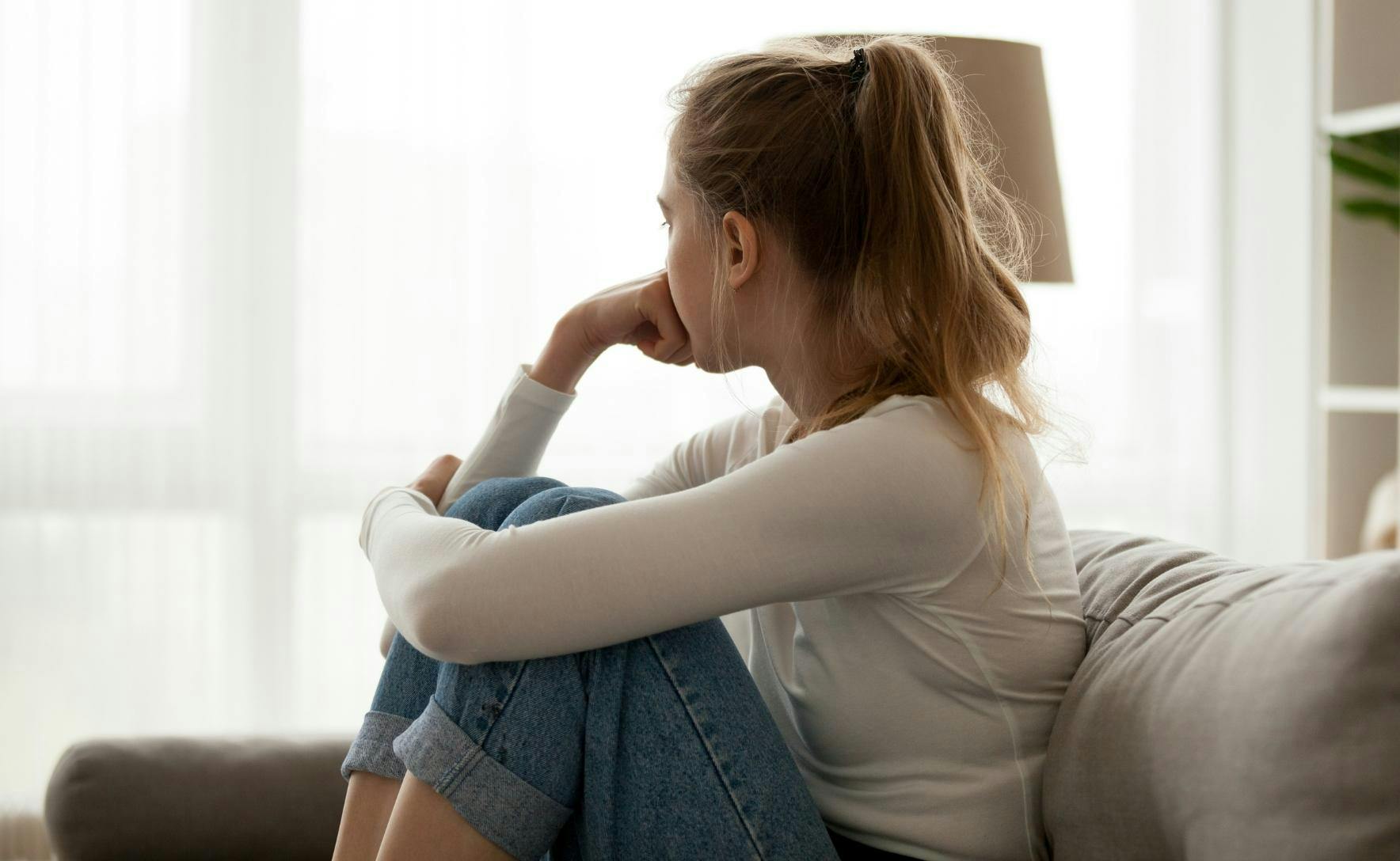 Side view young woman looking away at window sitting on couch at home