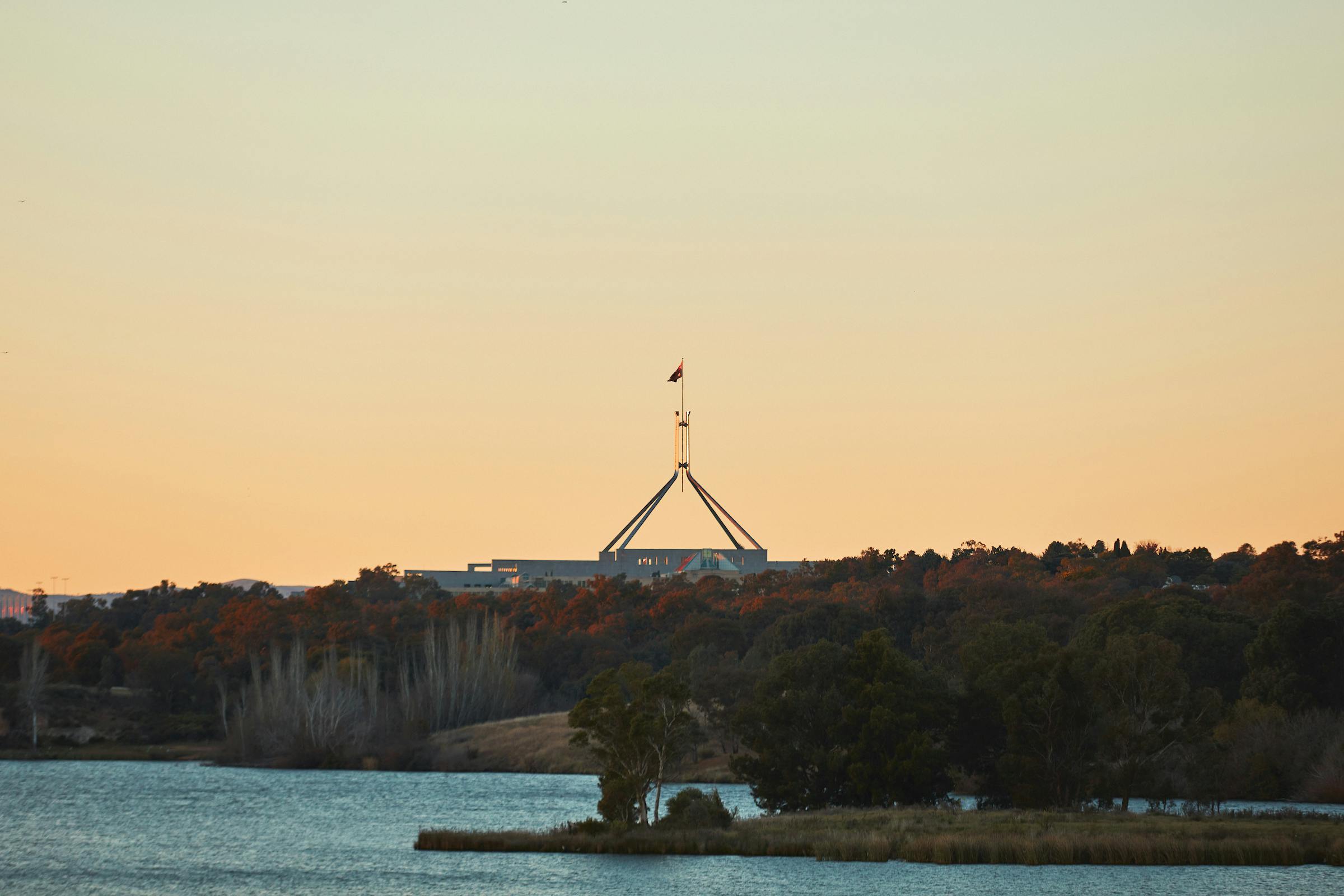 Parliament house with a flag flying on top is visible behind a lake at dusk