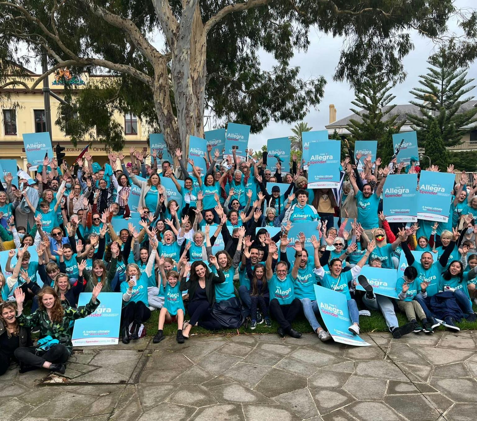 Group of people are pictured with their hands in the air celebrating. They are wearing teal coloured tshirts and holding teal coloured signs that read Allegra Spender