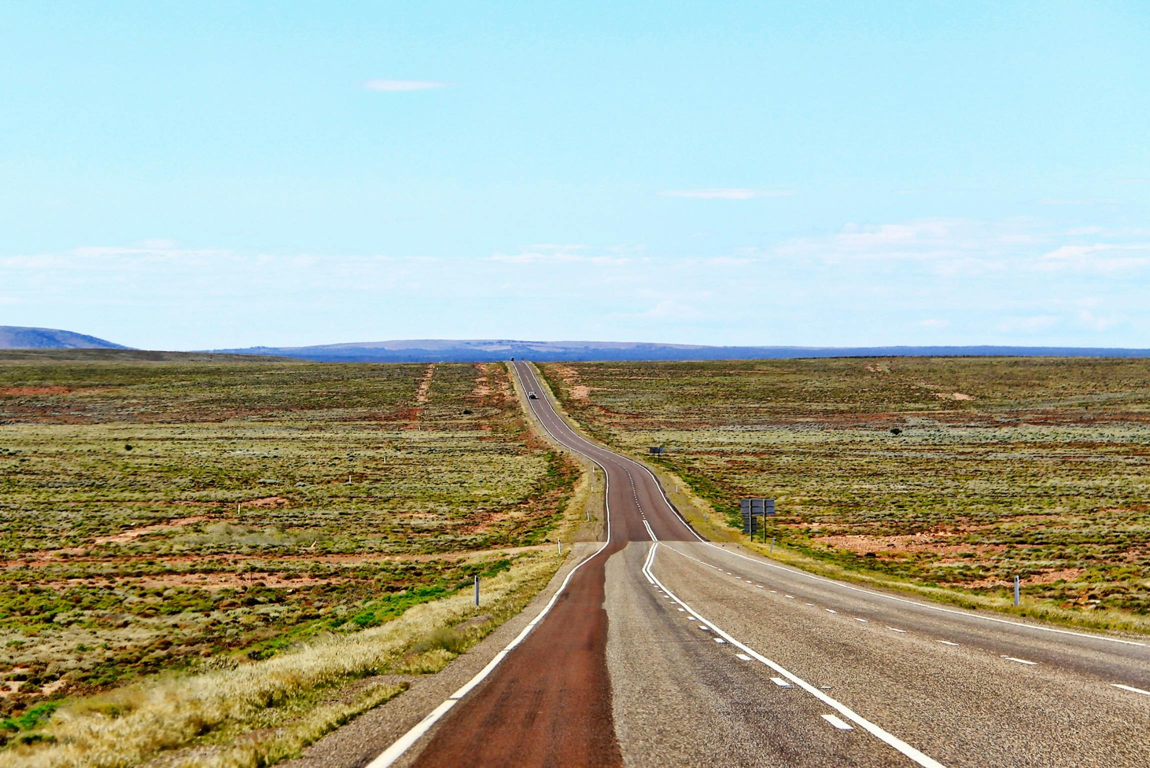 flat outback scene with grass covering the ground and low mountains in the background. A paved road cuts through the landscape