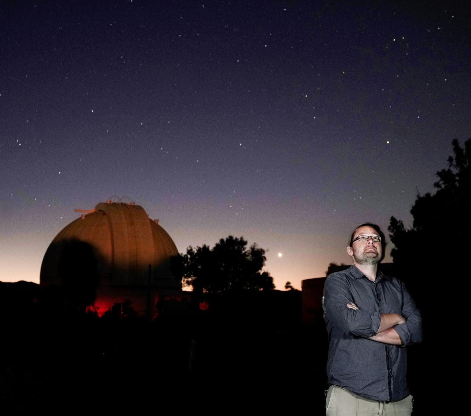 a man is standing withhis arms crossed looking at the night sky. In the background is a large round building and a starry night sky