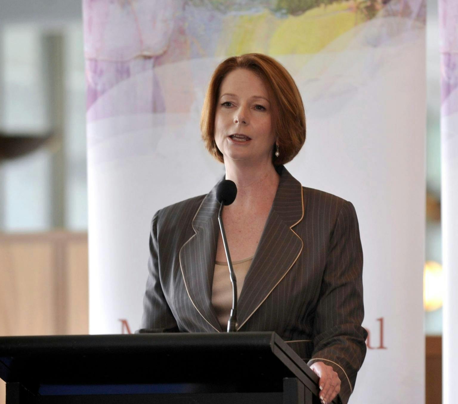 A woman is standing at a lecturn mid speech. She is wearing a grey suit and has red hair