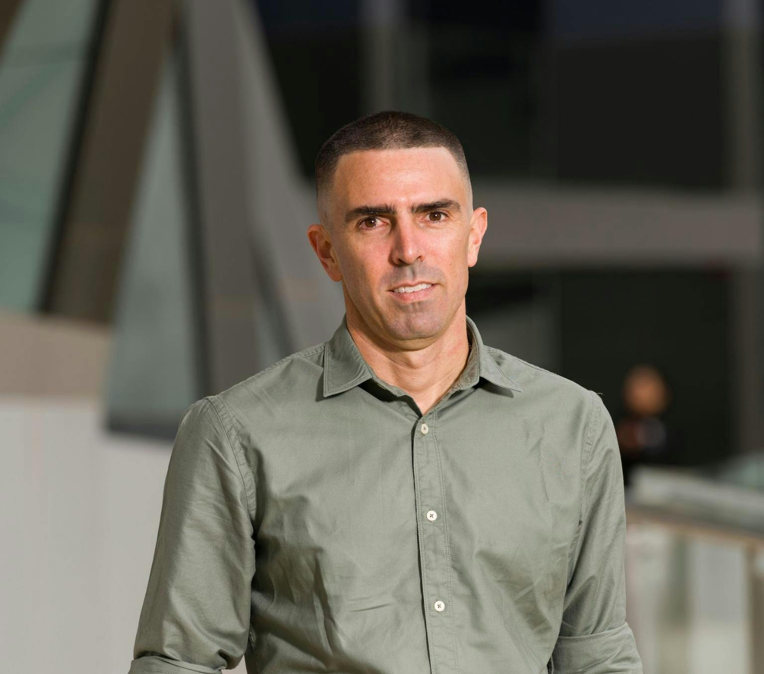 Jason is wearing a khaki coloured collared shirt. He is looking past the camera. Behind him is a blurred background of a building.