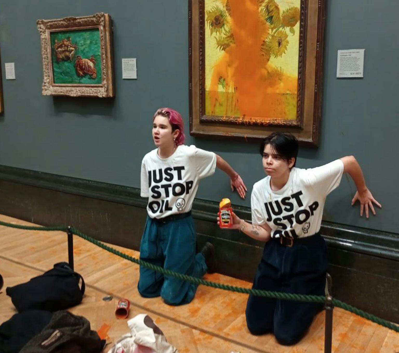 Two people in white shirts with the text 'JUST STOP OIL' kneel in front of a painting, and the palms of one hand each are placed against the wall. Red liquid is seen over the painting.
