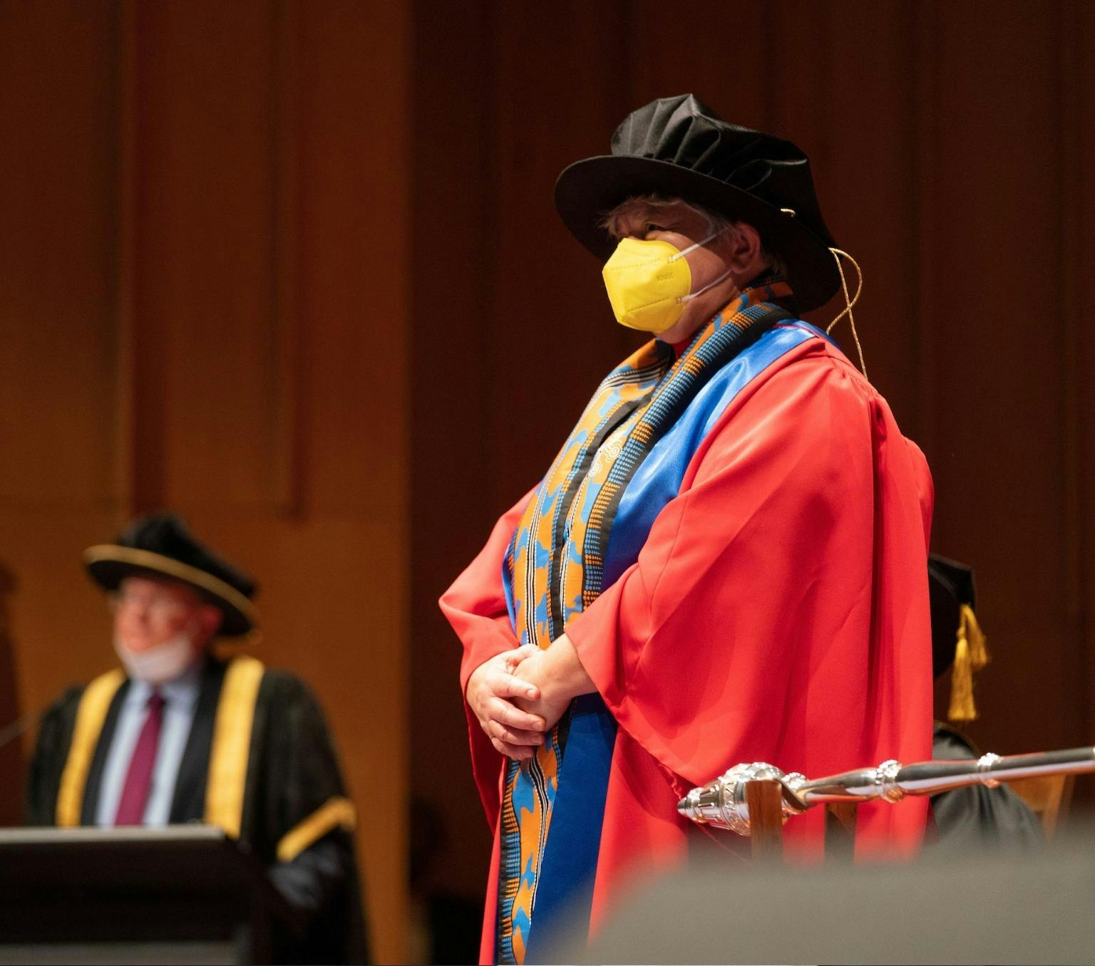 A woman is wearing academic regalia including a red and blue robe, and a large black hat. she is also wearing a yellow face mask