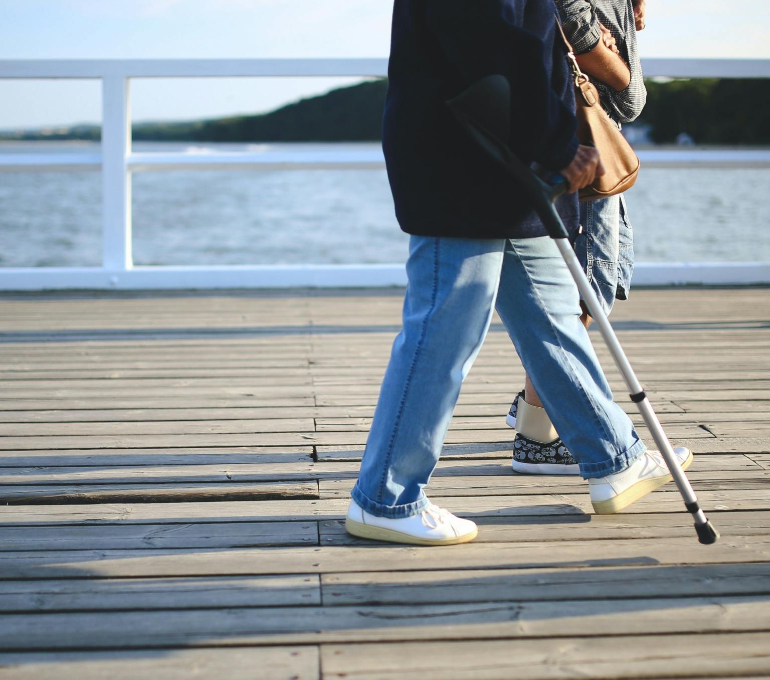 two people walking on a wooden pier over water. One person is using a walking stick