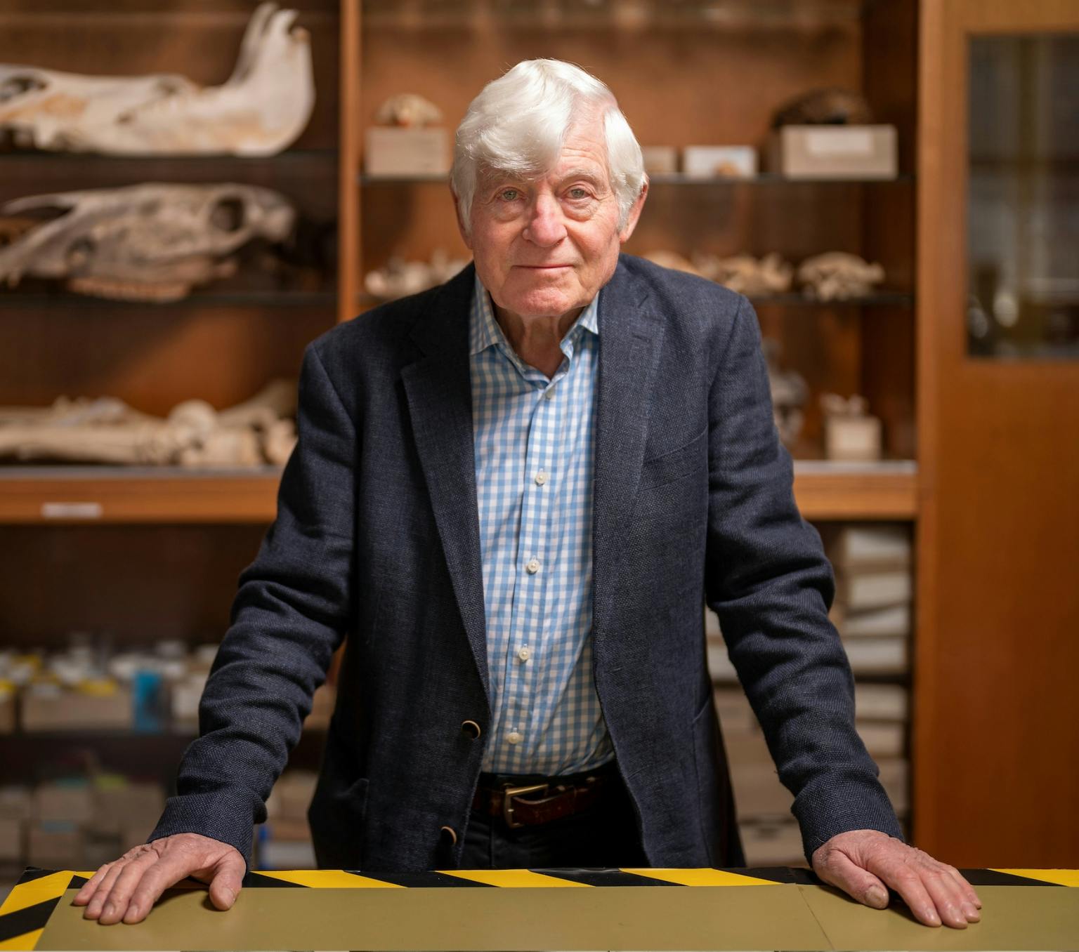 Peter is standing in front of a shelf with animal skulls and other artefacts. He has white short hair and s wearing a dark blue suit jacket
