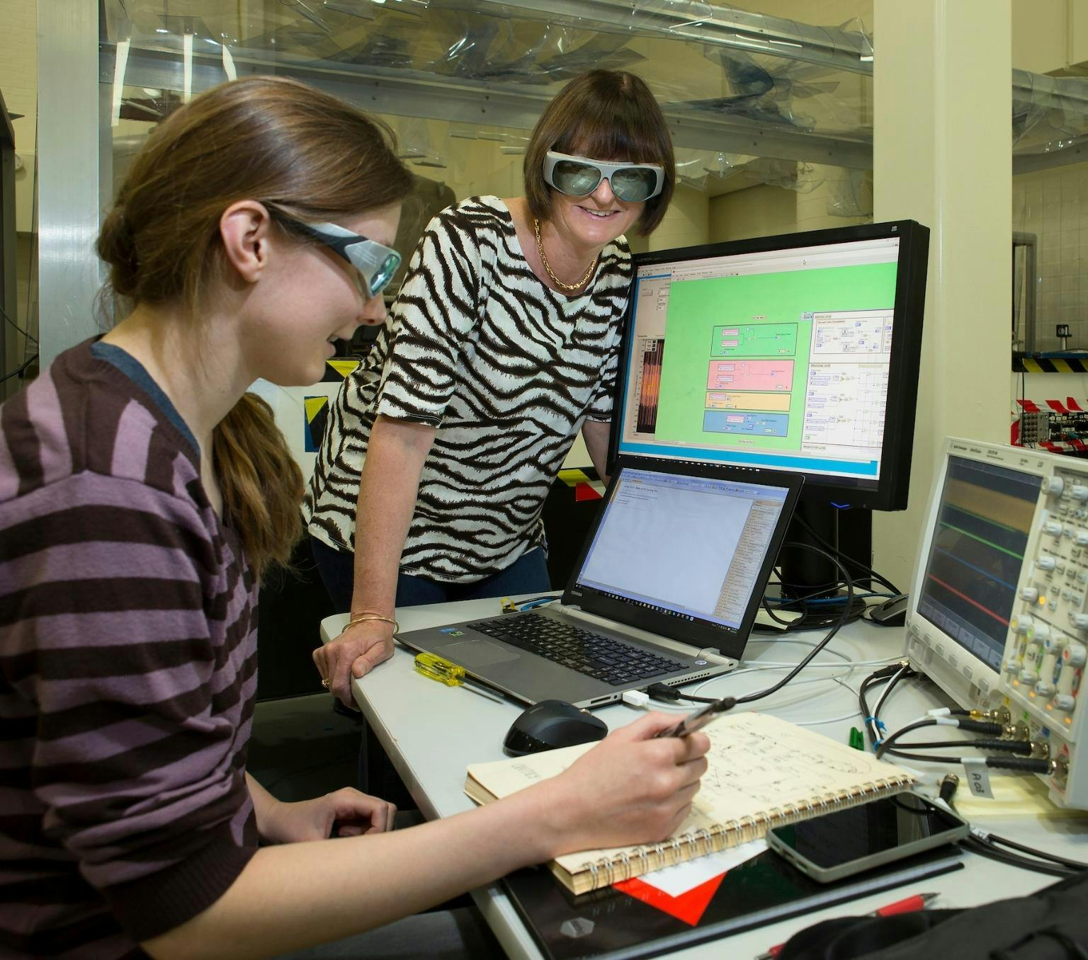 Susan is standing over a desk and watching a woman sitting at a computer with multiple screens around her. Both the women are smiling and wearing large grey reflective glasses