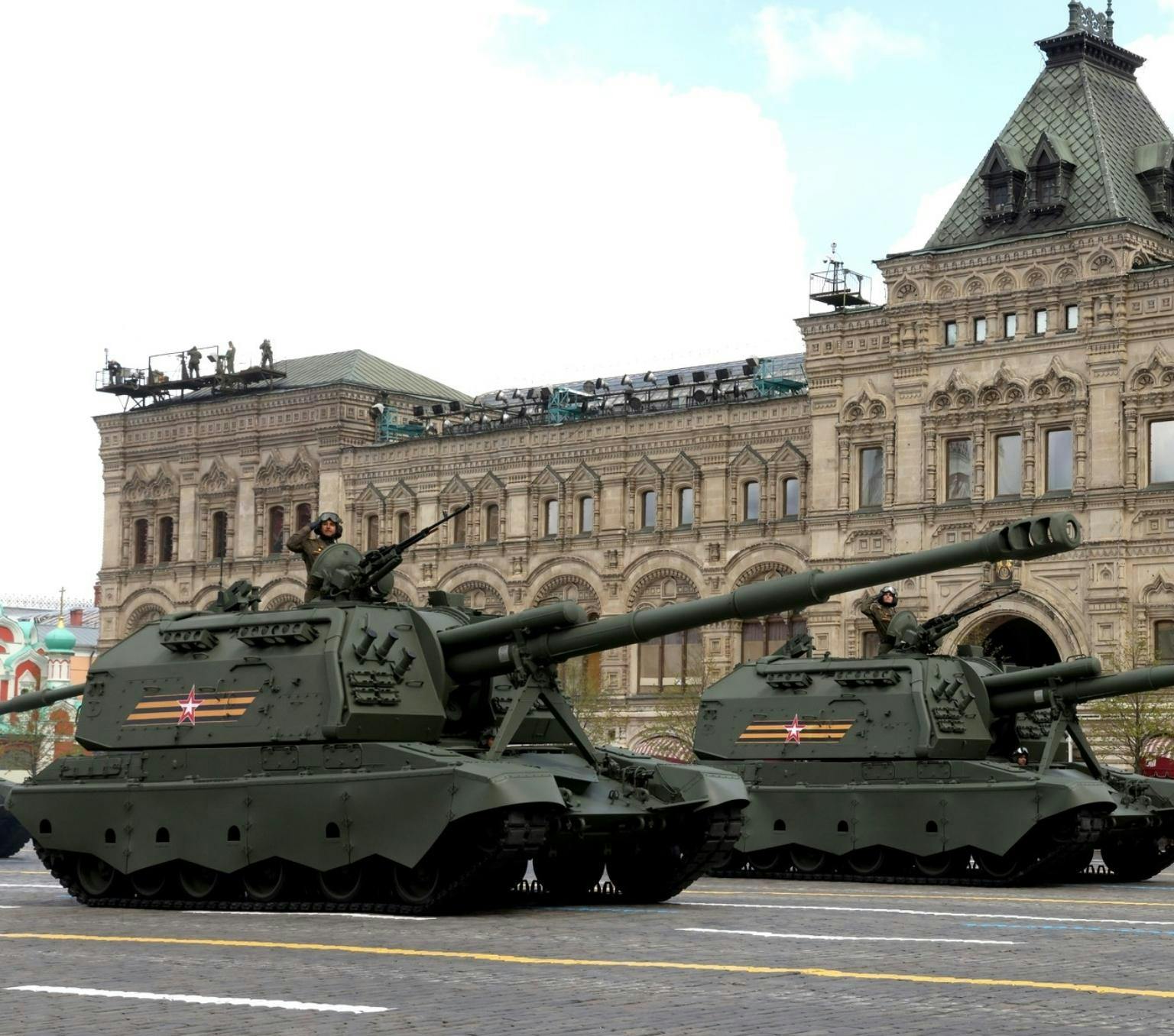 khaki coloured tanks are driving in front of a sandstone building