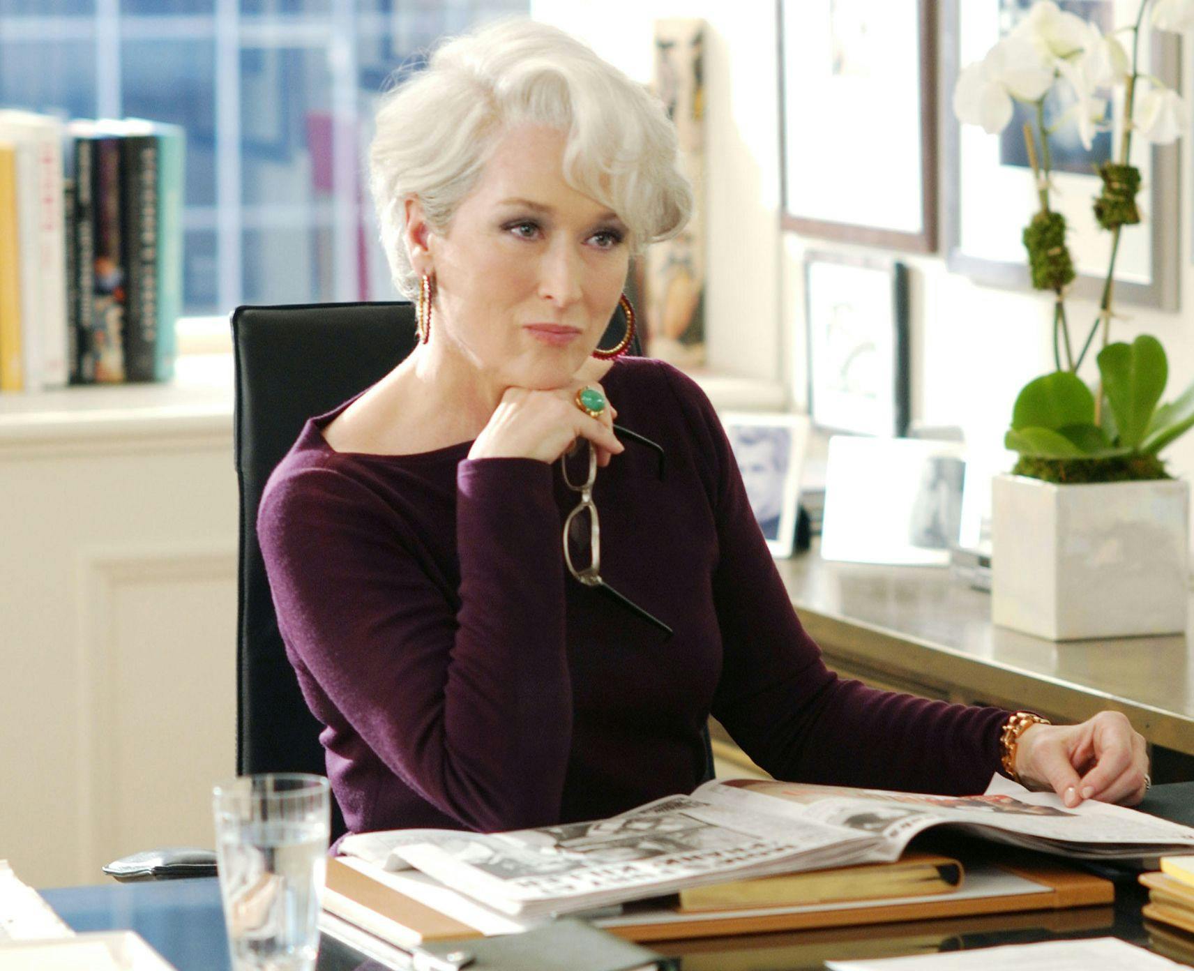 Meryl Streep as Miranda Priestly in the Devil wears Prada. She has a burgundy top on and is sitting at a desk looking unimpressed.