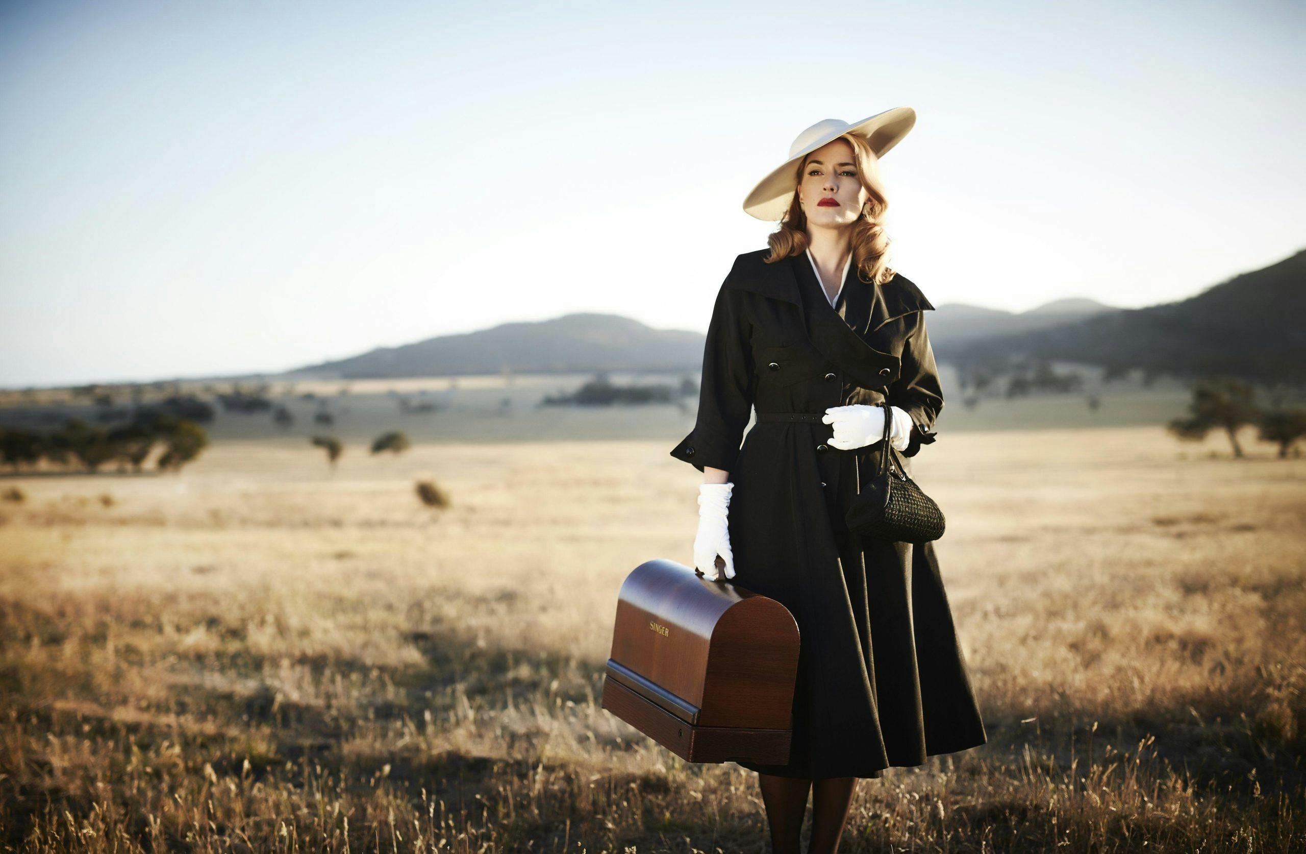 Kate Winslet is dressed in costume as Tilly Dunnage, in a black vintage dress, with white gloves and a white hat. She is standing in front of a field of dry coloured grass, holding a suitcase.