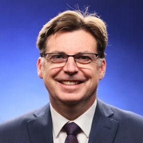 A man with brown hair and glasses smiles at the camera. He is wearing a brown tie and a dark grey suit jacket.