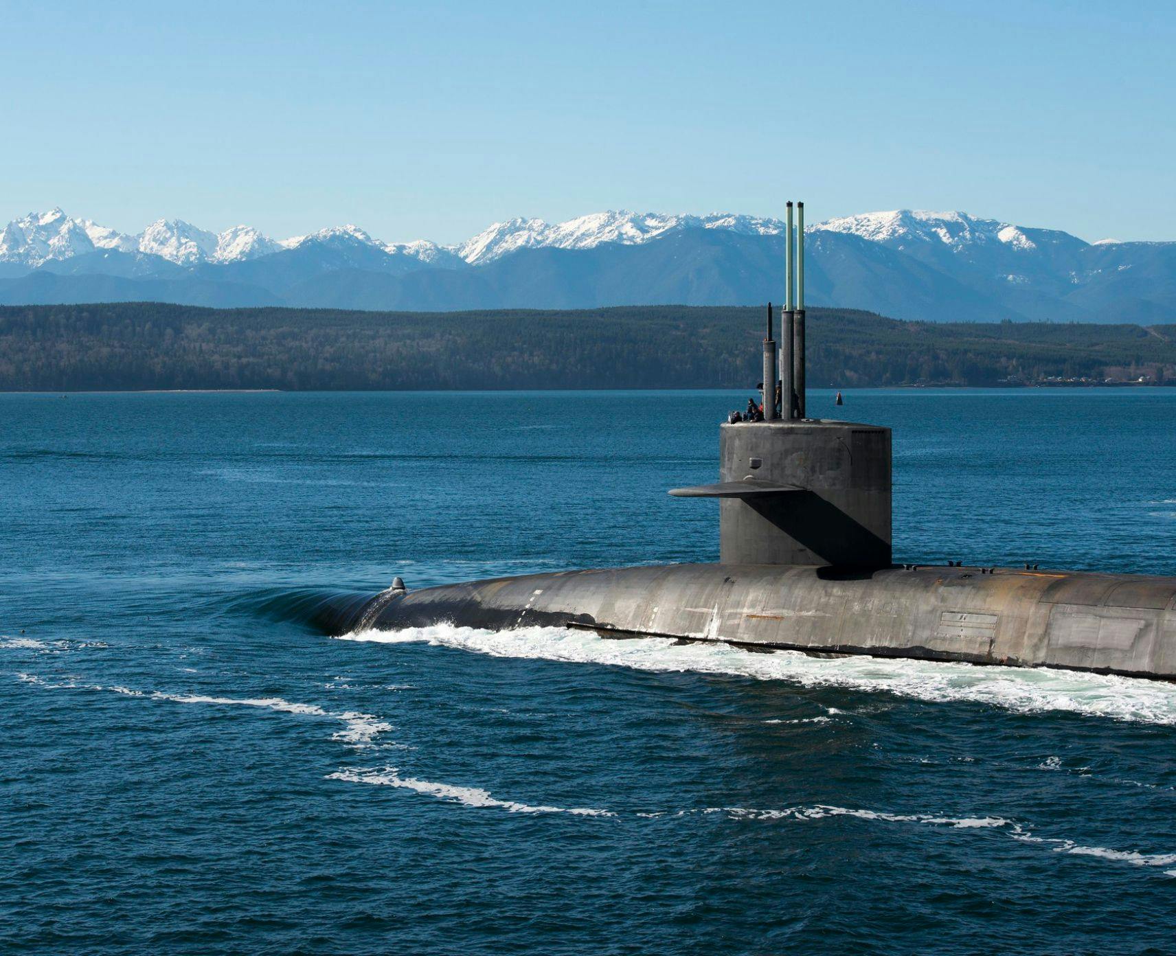 United States submarine returning to port with snow-capped mountains in the background.