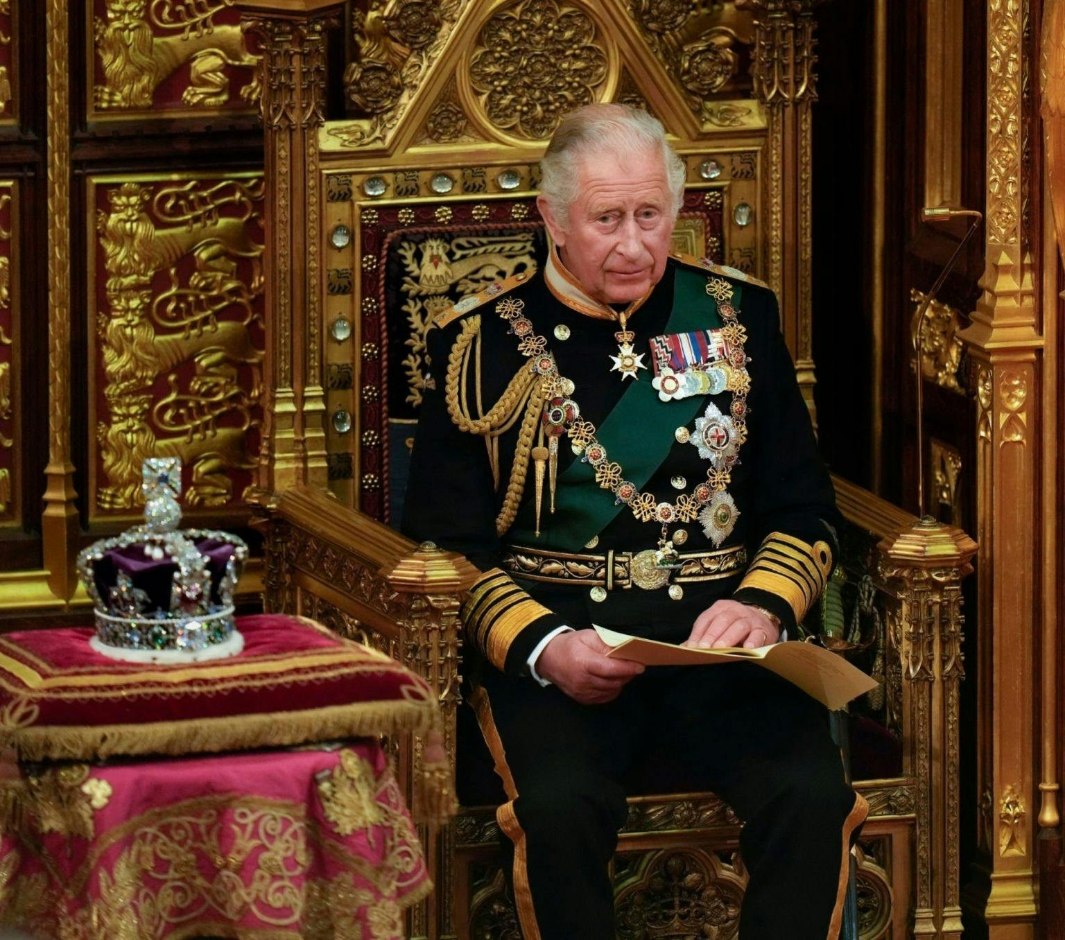 King Charles sits on a throne. A crown sit on the table to his right.