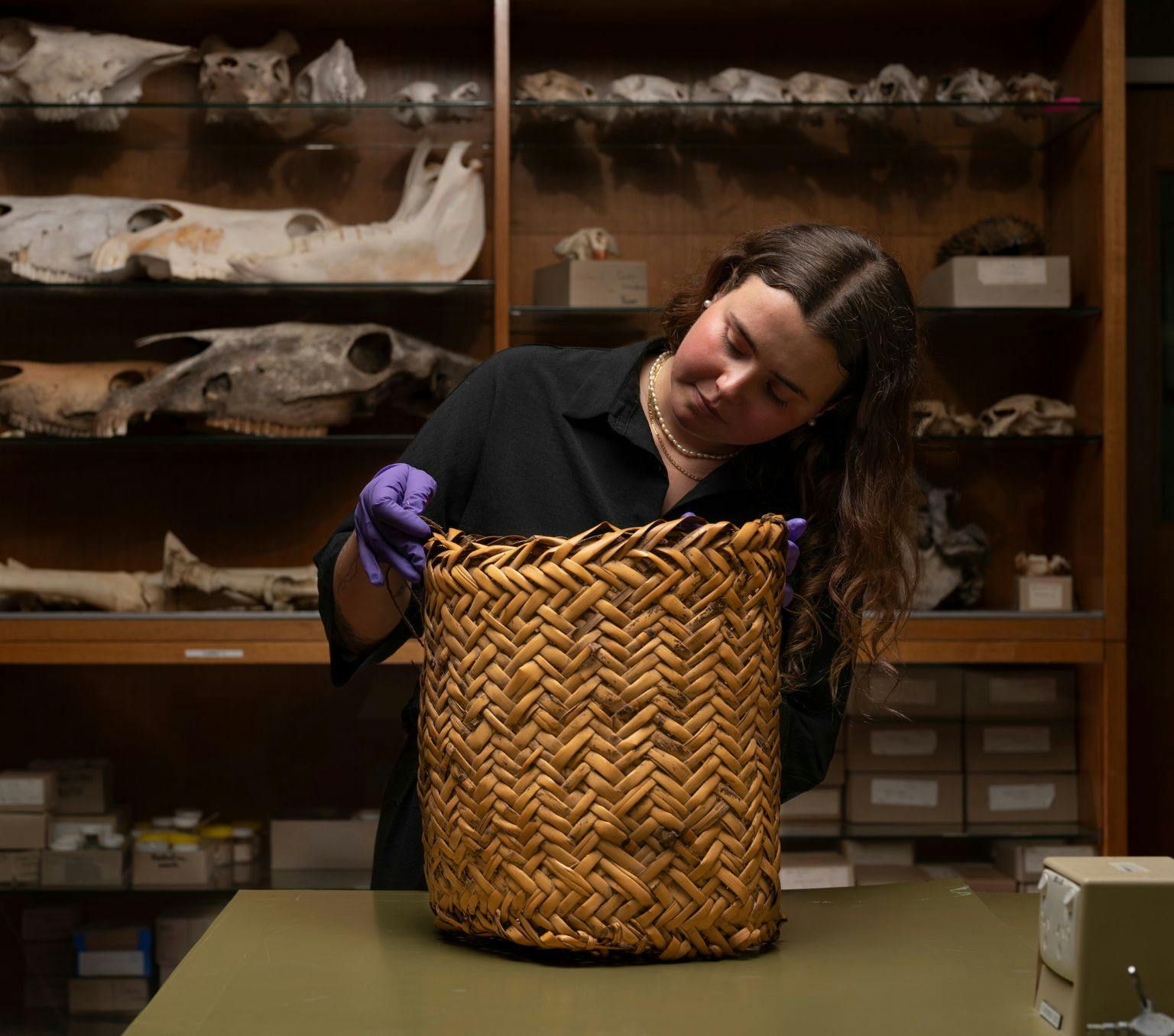 A woman wearing purple gloves gently touchers a large woven basket. On the shelves behind her are all sorts of objects including bones.