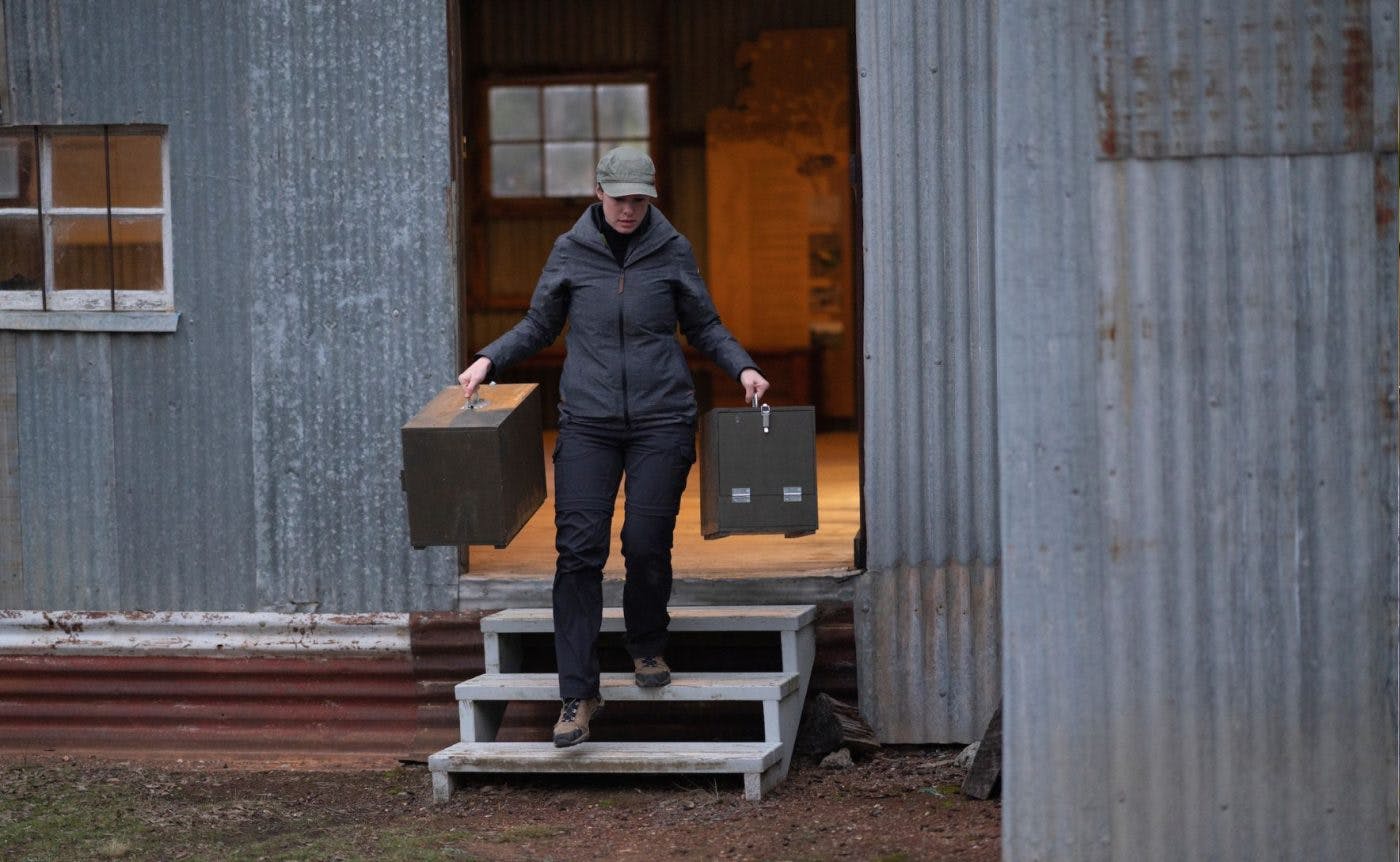 Belinda Wilson descends a set of stairs from a shed made out of corrugated iron. She is carrying a large square case in each hand and is dressed in dark clothes and a khaki cap.