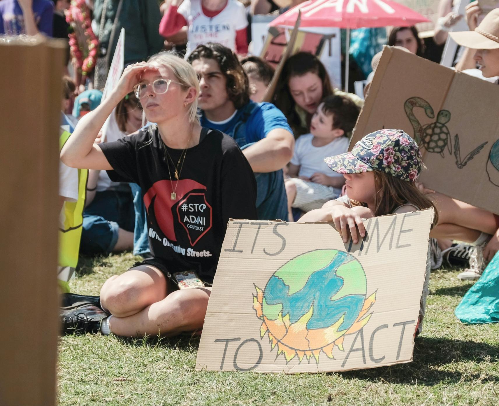 Two young protestors sit down on grass. Their sign reads its time to act.